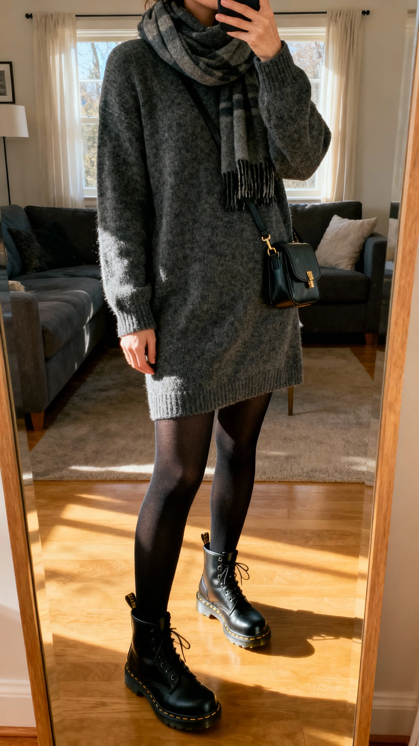iPhone living-room mirror selfie of a woman wearing a charcoal sweater dress with black tights, black Doc Martens, scarf, and small shoulder bag, face not visible, cropped at neck, soft afternoon window light, natural iPhone photo quality.