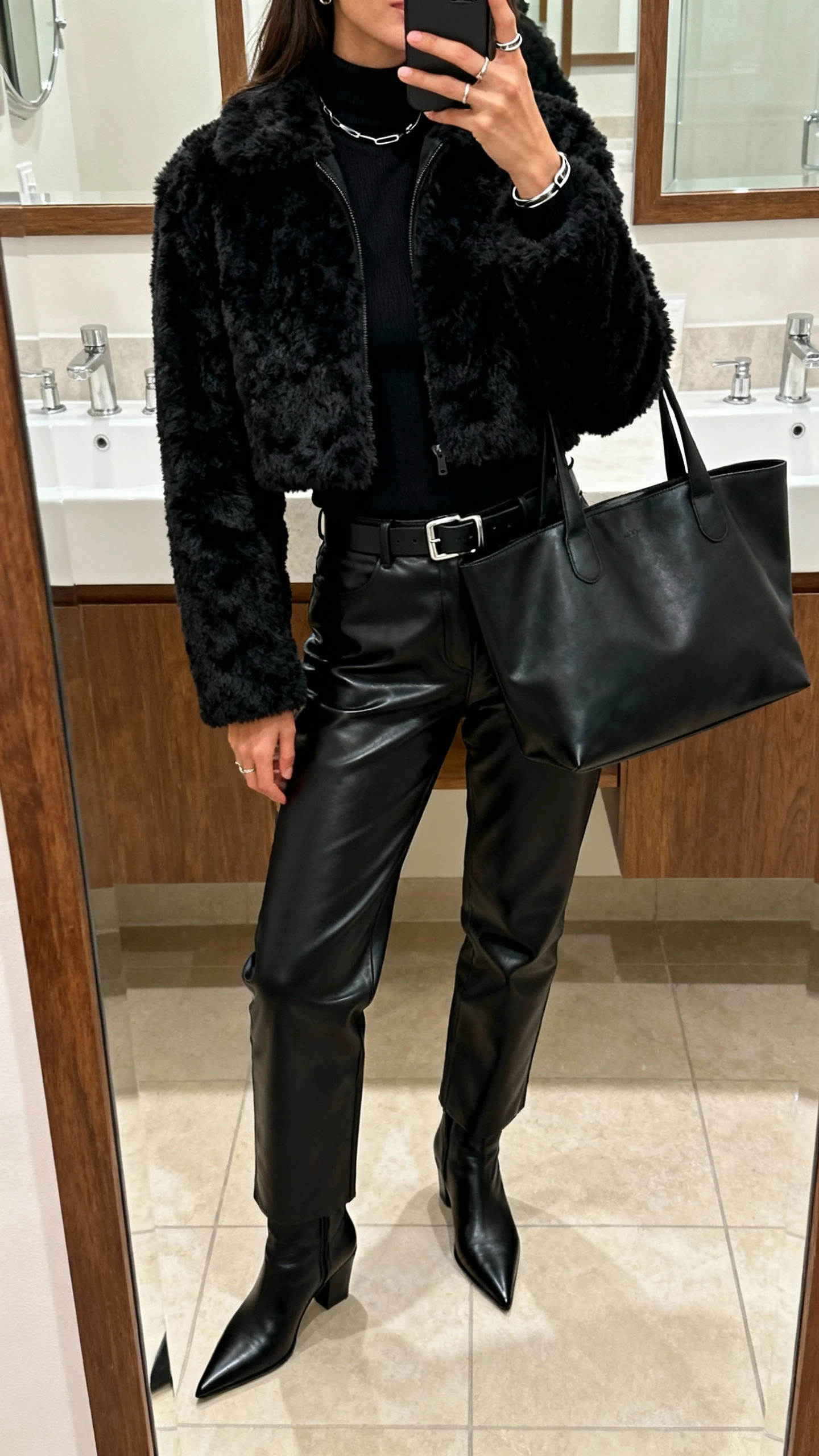 iPhone mirror selfie cropped at neck showing a woman in an all-black look: black shaggy cropped faux fur jacket, black mock-neck top, black leather trousers, pointed-toe black ankle boots, oversized black tote, minimalist gunmetal jewelry, black belt with subtle buckle; bathroom mirror, soft indoor lighting, natural iPhone photo quality.