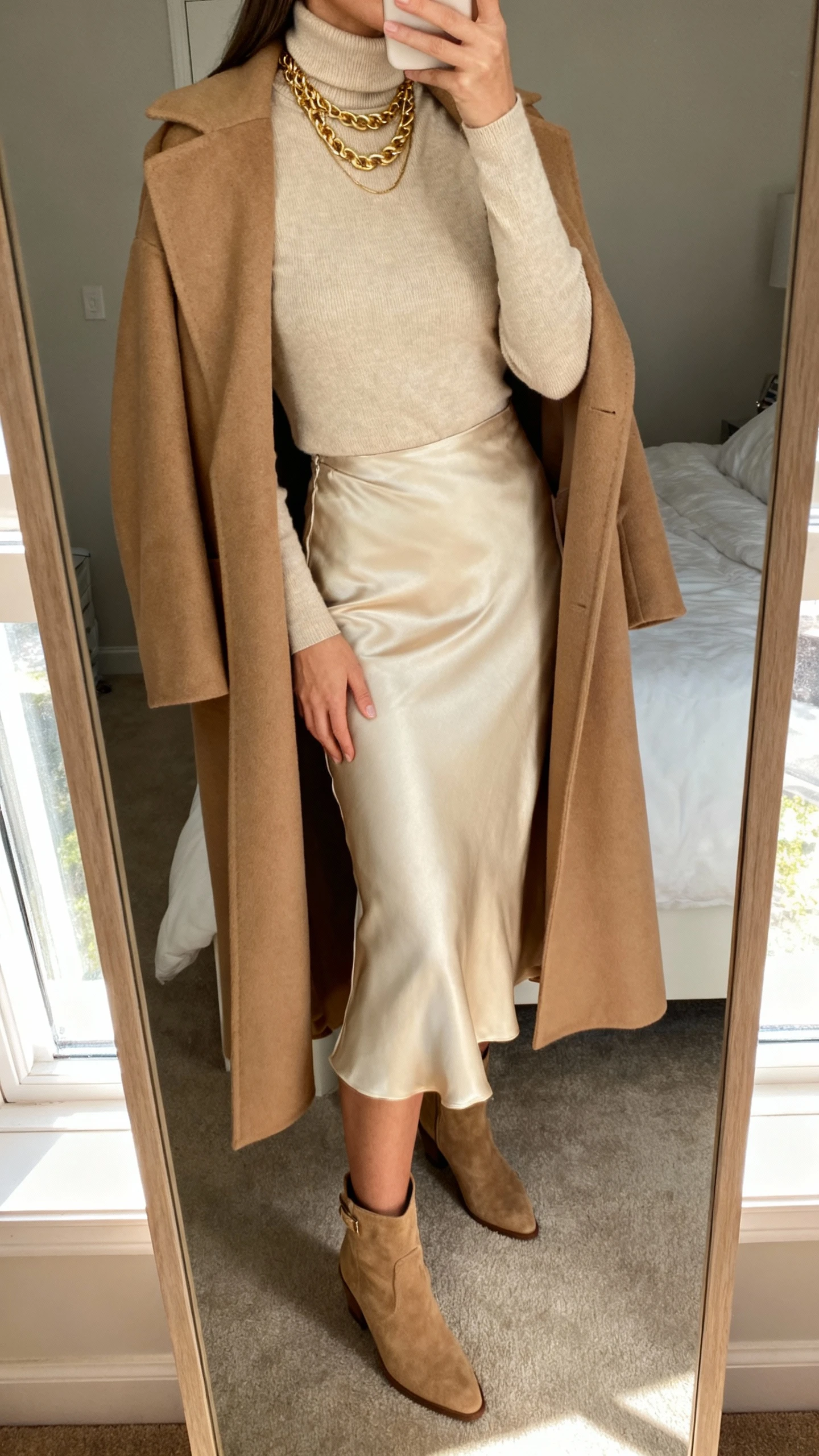 iPhone mirror selfie cropped at neck showing a woman in a beige turtleneck, champagne slip skirt, long camel coat draped over shoulders, tan ankle boots, layered gold chains, face not visible, bedroom mirror with window light, natural iPhone quality.