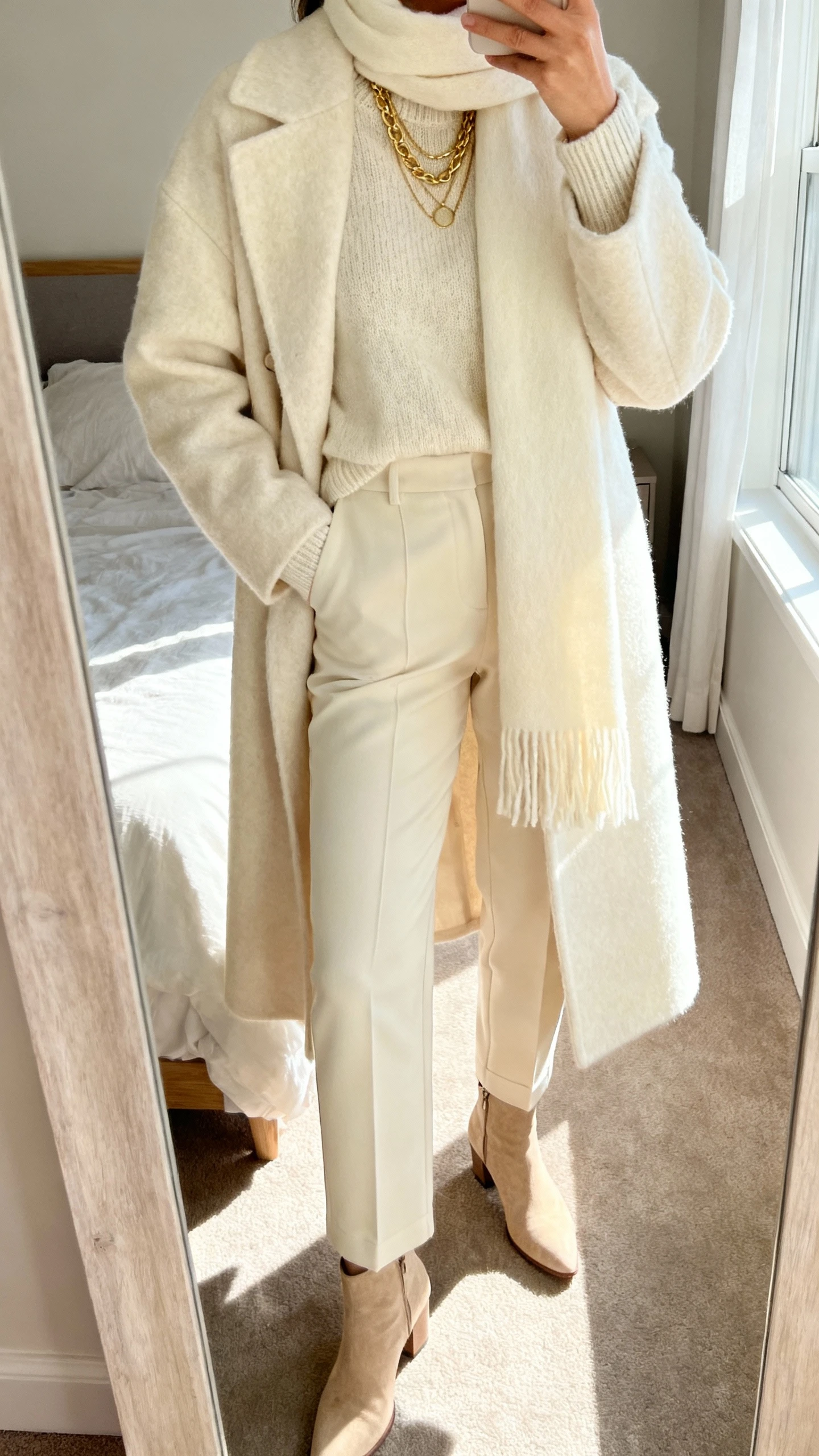 iPhone mirror selfie cropped at shoulders showing a woman in head-to-toe cream: wool coat, cashmere sweater, tailored trousers, cream scarf, beige ankle boots, gold layered necklaces, face not visible, bedroom mirror with morning window light, natural iPhone photo quality.