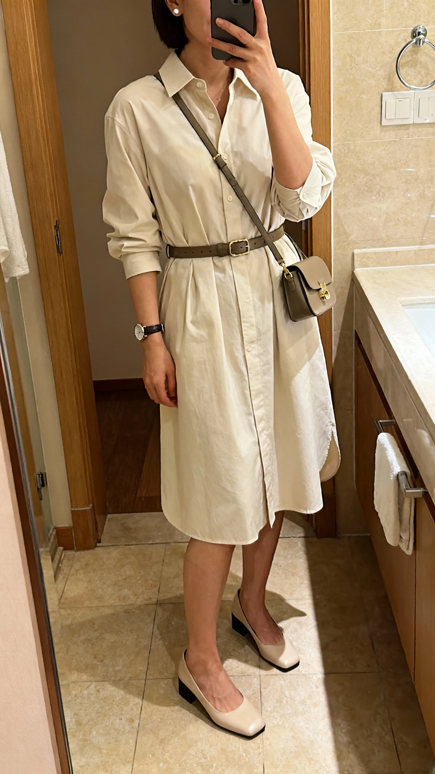 iPhone mirror selfie of a woman in a classic shirt dress cinched with a belt, simple studs, wristwatch, low block heels, small crossbody bag, face not visible, bathroom mirror, warm indoor lighting, natural iPhone photo quality.