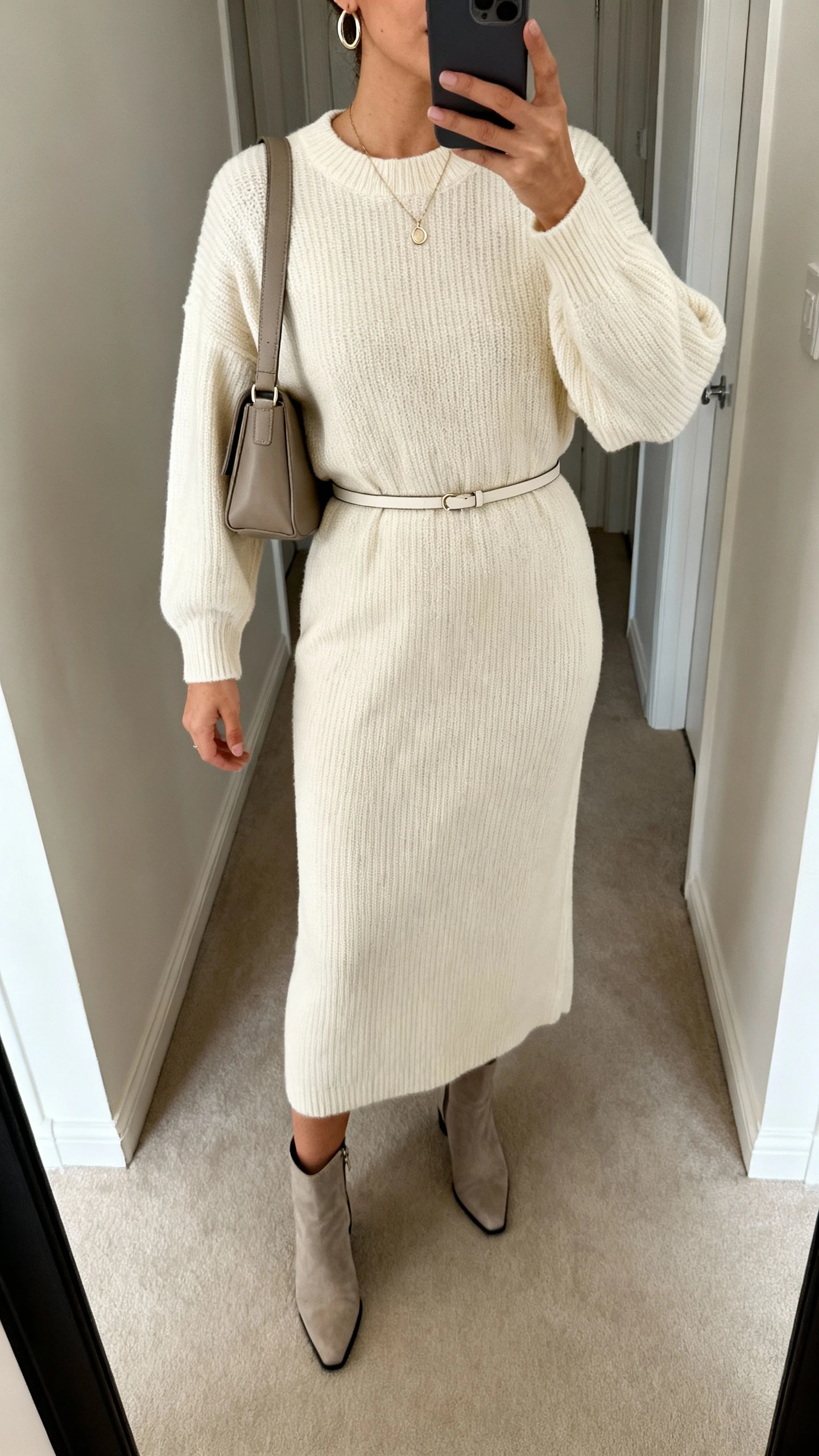 iPhone mirror selfie of a woman in a cream knit midi dress with thin belt, sleek ankle boots, simple hoop earrings and a dainty pendant, structured shoulder bag; cropped at neck, hallway mirror, soft indoor lighting, face not visible, natural iPhone photo quality.