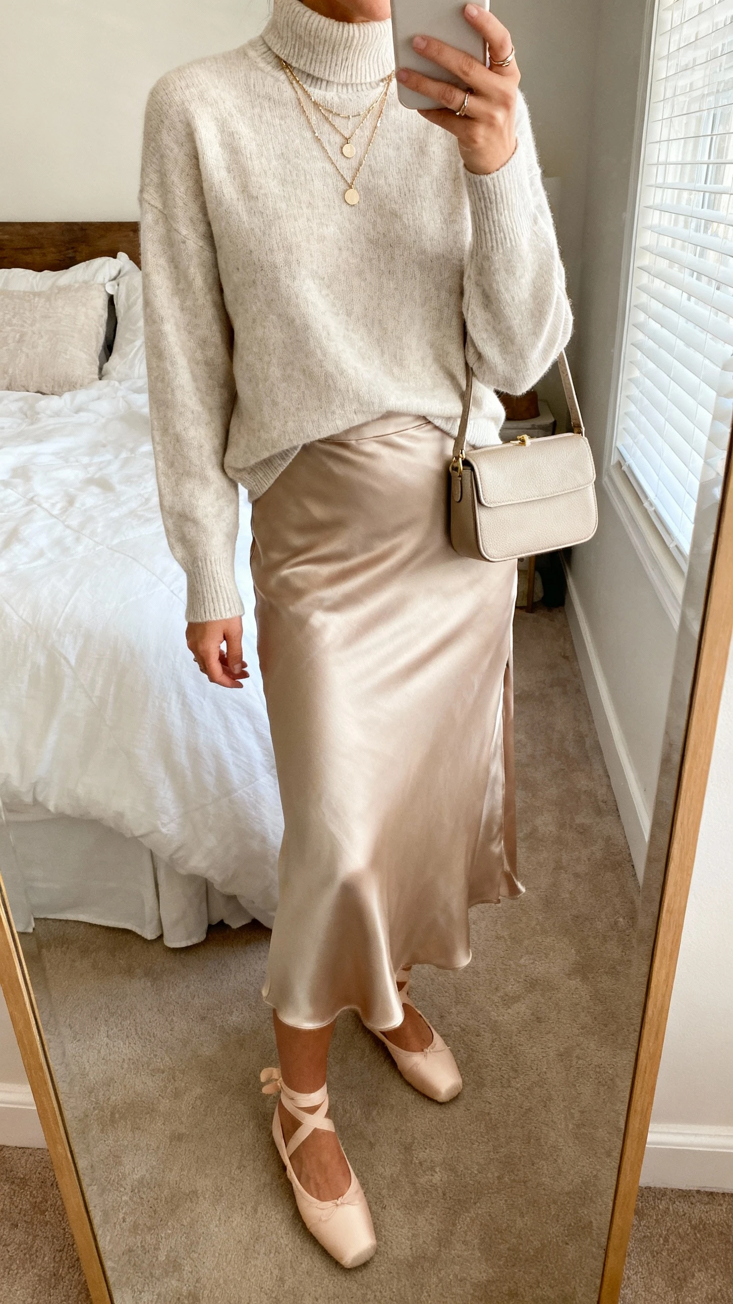 iPhone mirror selfie of a woman in a lightweight mock-neck sweater tucked into a satin bias-cut midi slip skirt, refined ballet flats, layered delicate necklaces, structured mini bag; cropped at neck, soft bedroom light, face not visible, natural iPhone photo quality.