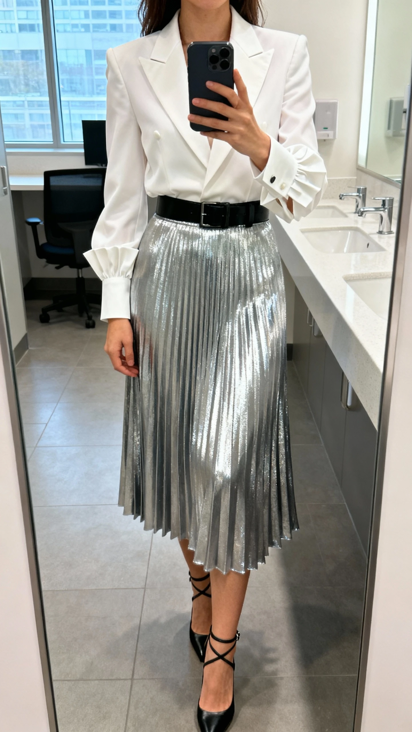 iPhone mirror selfie of a woman in a metallic pleated midi with a sculptural tux shirt: silver pleated midi skirt, crisp white tuxedo-style shirt with structured cuffs and collar, black belt, ankle-strap heels; face not visible, office restroom mirror, bright indoor light, natural iPhone quality.