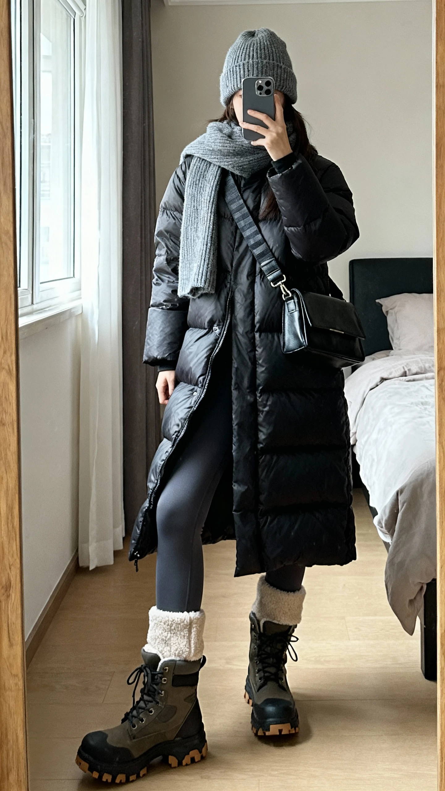 iPhone mirror selfie of a woman in a mid-length matte black puffer coat, high-rise fleece-lined leggings, chunky waterproof lug-sole boots, cashmere beanie with matching scarf, and a structured crossbody with thick strap; bedroom mirror, natural window light, face not visible, natural iPhone photo quality.
