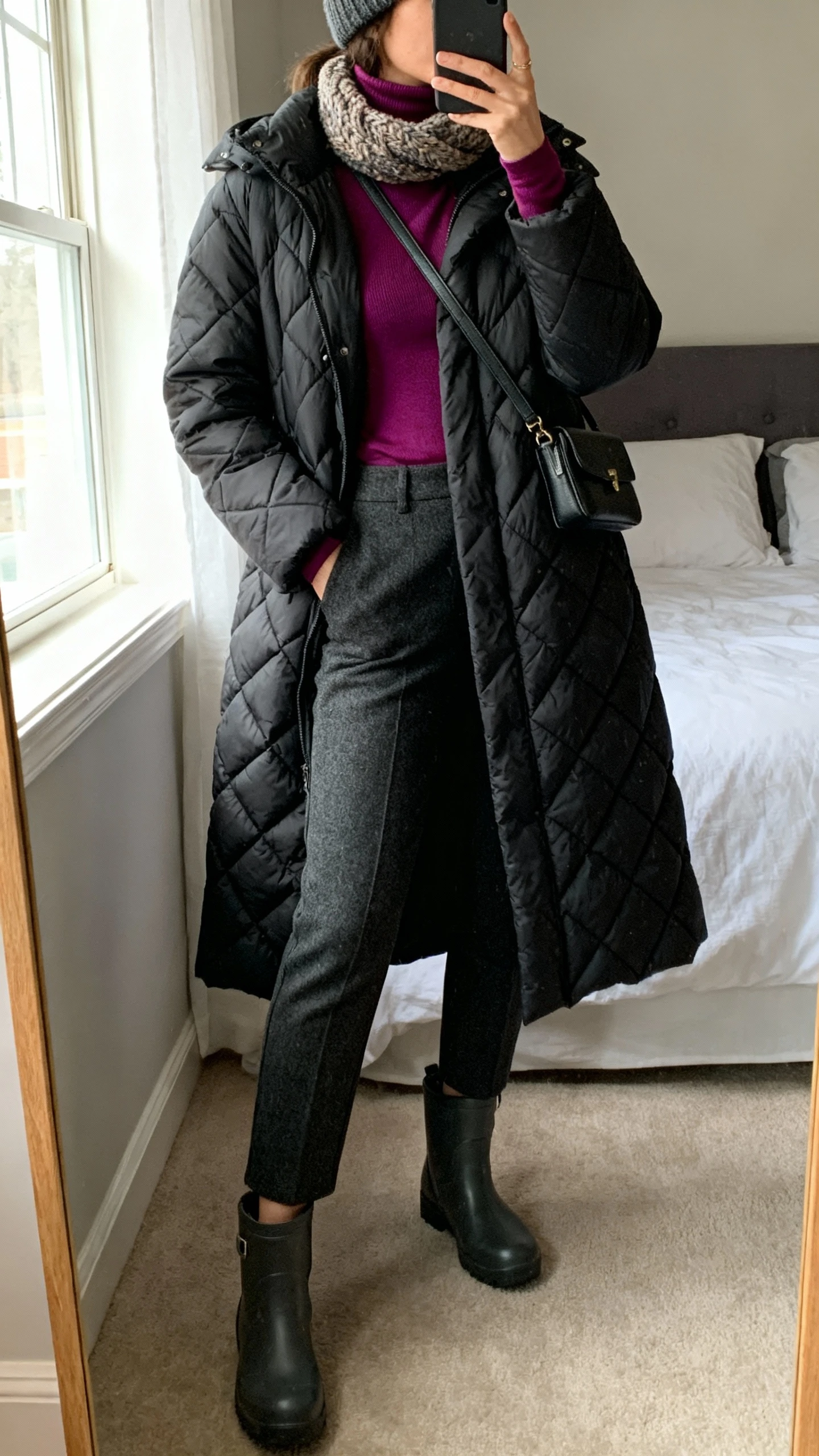 iPhone mirror selfie of a woman in a mid-length quilted puffer coat (black), fitted jewel-tone turtleneck, slim wool trousers, waterproof ankle boots, chunky scarf, beanie, and a crossbody bag; bedroom full-length mirror, cropped at neck, face not visible, window light, natural iPhone photo quality.