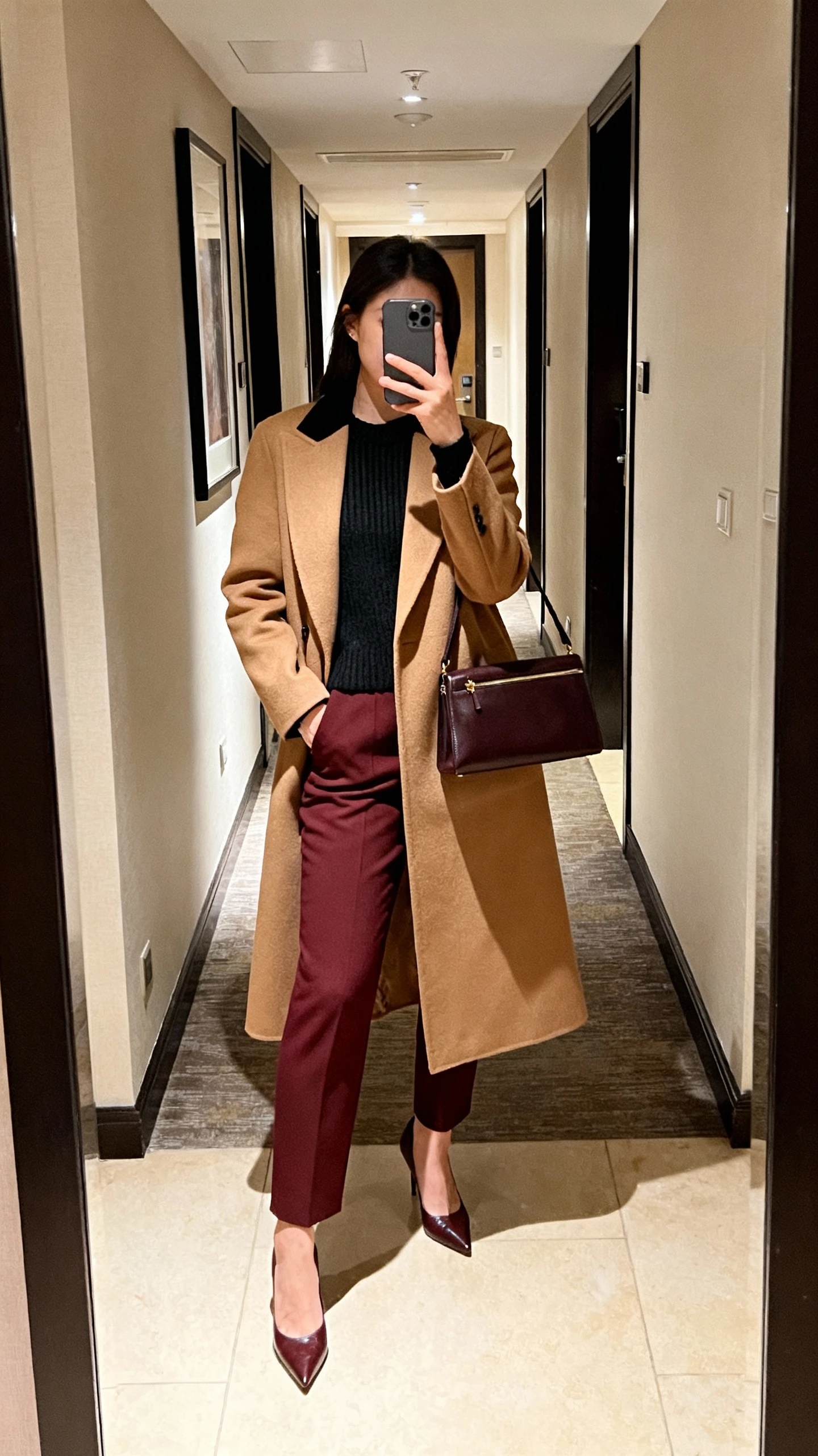 iPhone mirror selfie of a woman in a monochrome outfit head-to-toe camel (or black, or burgundy): tailored coat, knit, matching trousers, tonal heels, structured bag, face not visible, hotel hallway mirror, ambient light, casual iPhone aesthetic.