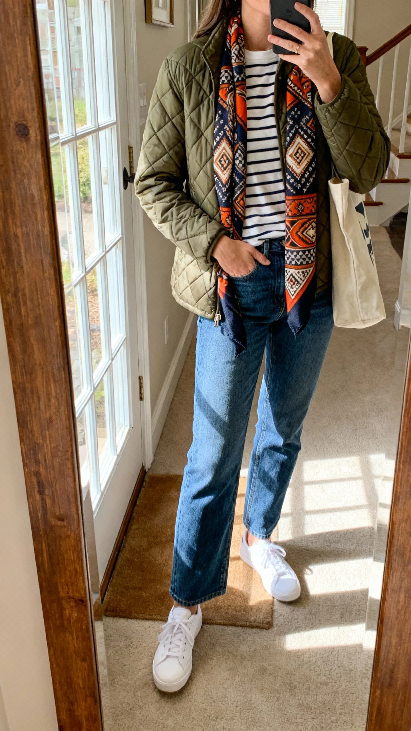 iPhone mirror selfie of a woman in a quilted olive jacket over a striped tee, straight-leg blue jeans, white sneakers, bold patterned statement scarf, tote bag, face not visible, entryway mirror, afternoon window light, natural iPhone photo quality.