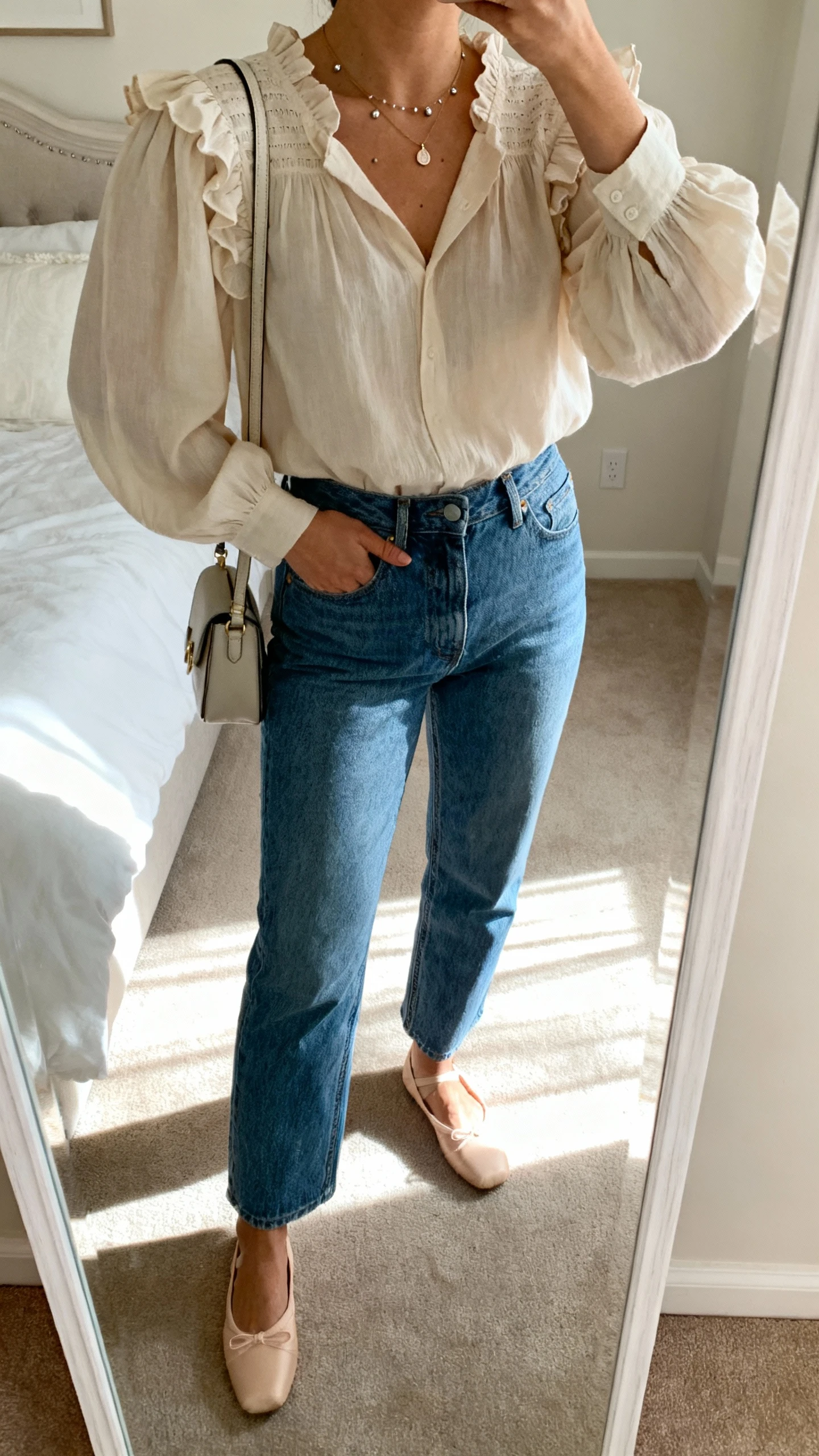 iPhone mirror selfie of a woman in a romantic blouse (soft ruffles or puff sleeves) tucked into blue jeans, ballet flats, dainty pendant and studs, small shoulder bag, face not visible, cropped at neck, bedroom mirror with morning light, natural iPhone quality.