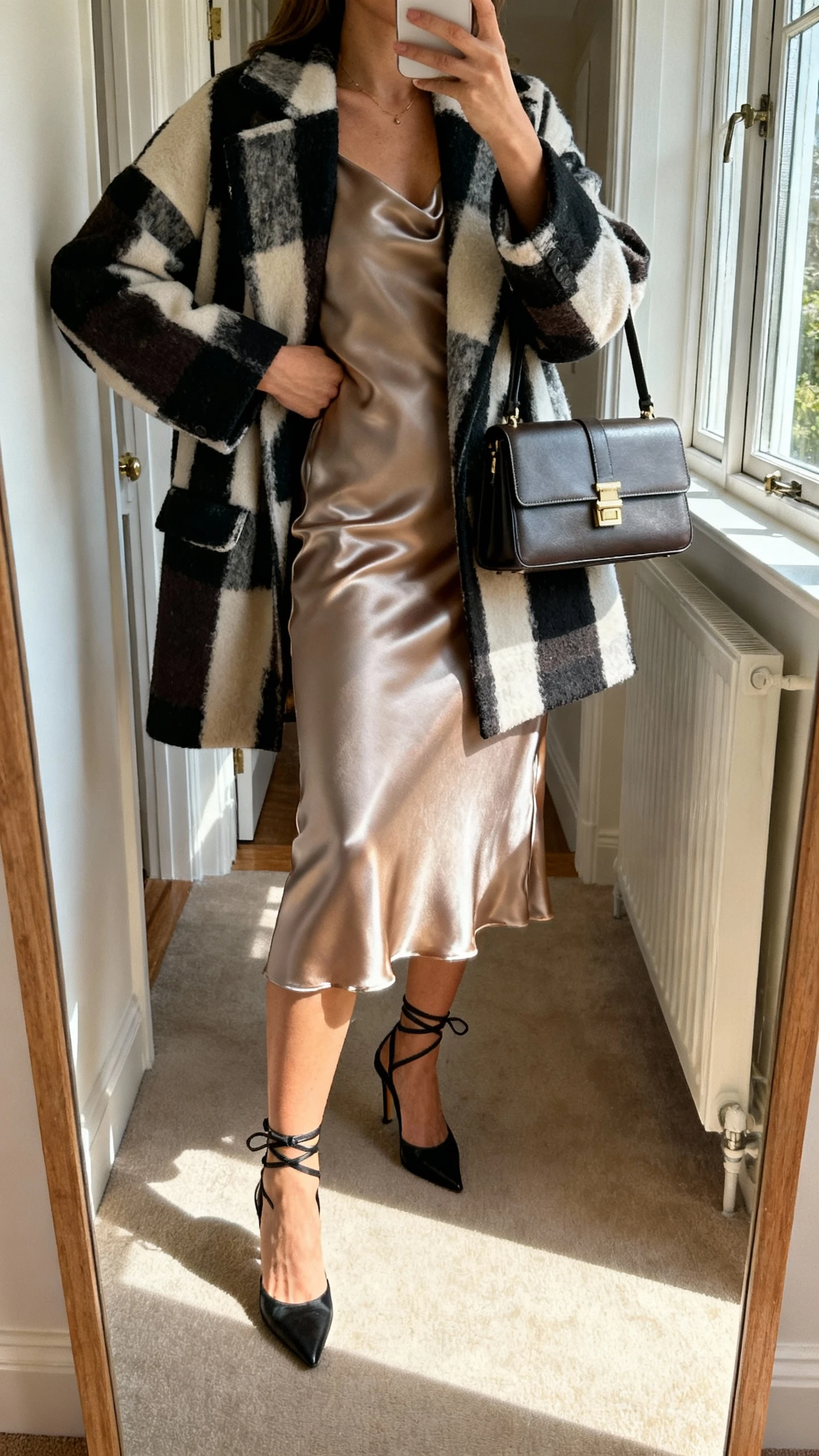 iPhone mirror selfie of a woman in a satin midi dress topped with a bold statement coat, ankle-strap heels and structured handbag, face not visible, entryway mirror, morning light from window, natural iPhone photo quality.