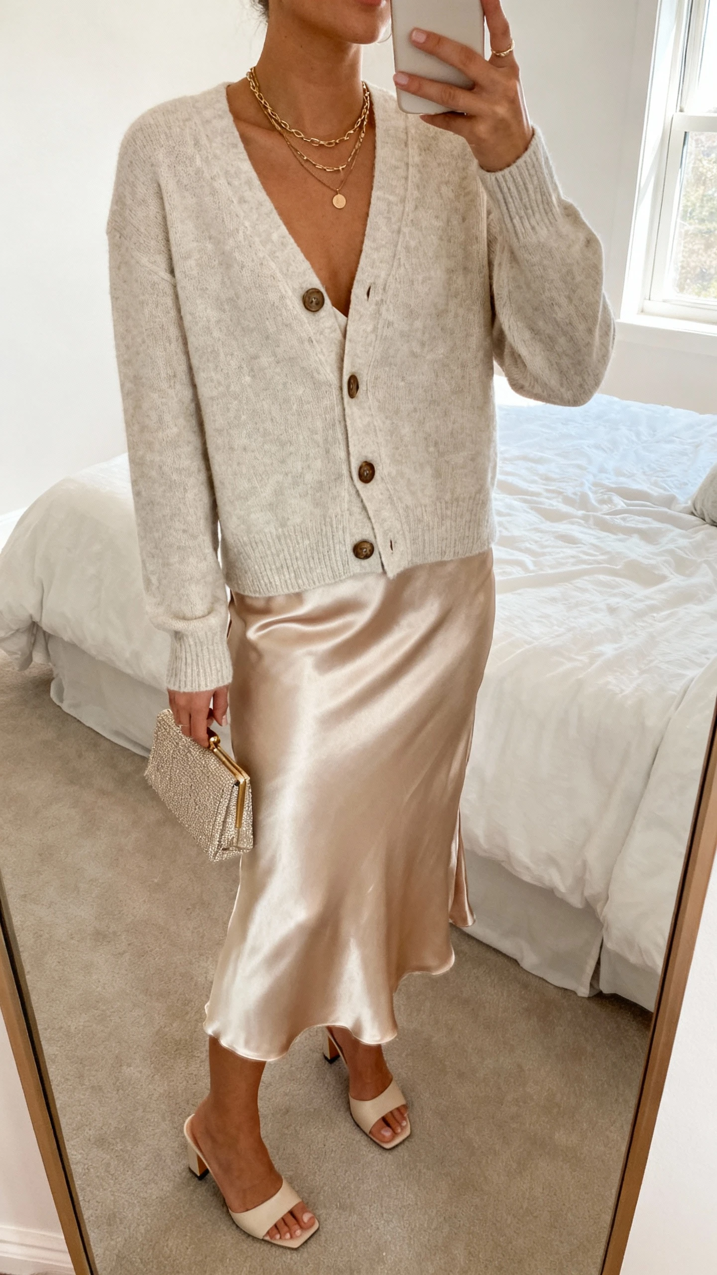 iPhone mirror selfie of a woman in a soft cardigan slightly unbuttoned over a satin slip skirt, delicate layered necklaces, kitten-heel mules, small clutch; face not visible, cropped at neck, bedroom mirror with gentle window light, natural iPhone photo quality