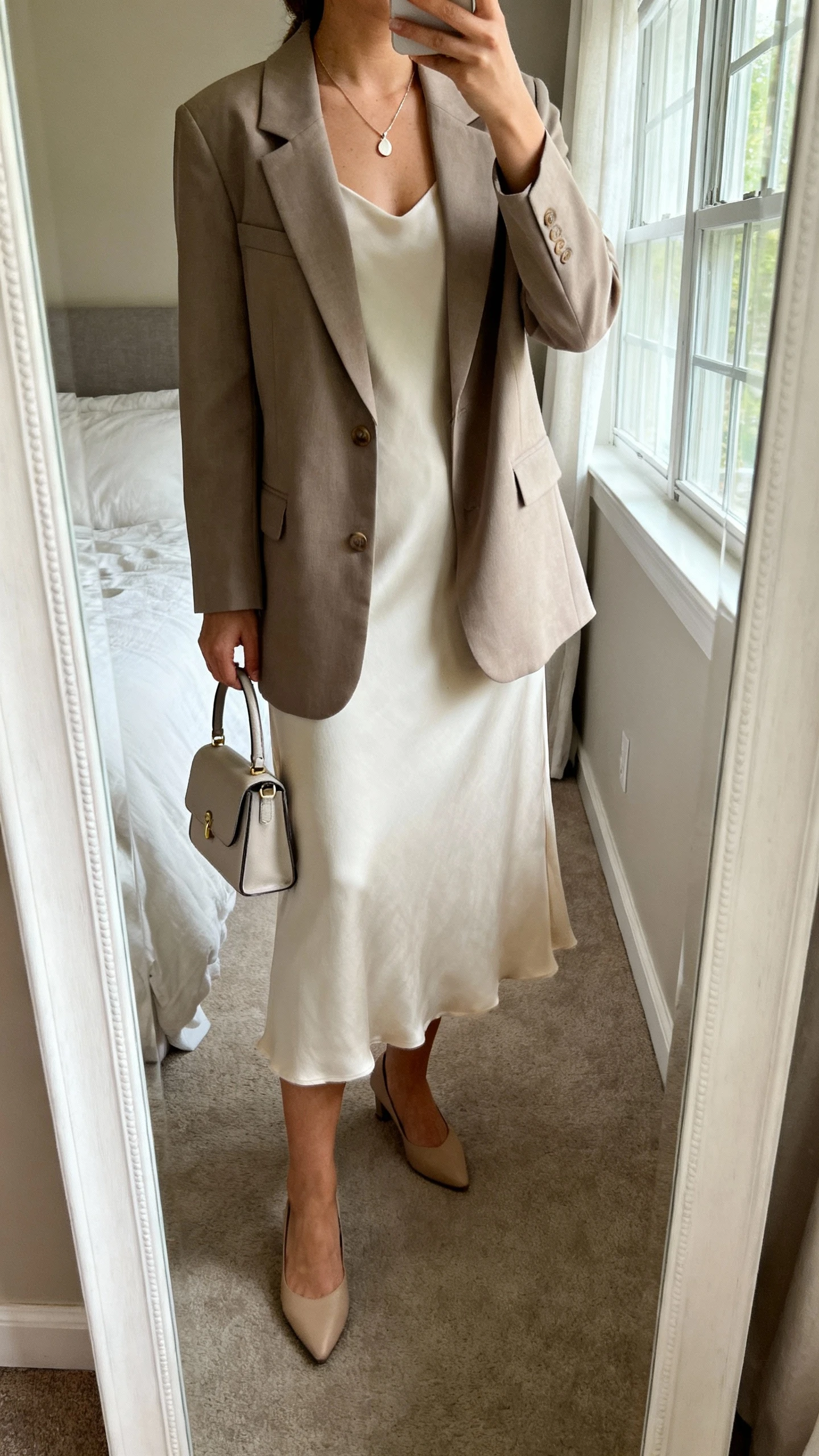 iPhone mirror selfie of a woman in a soft midi dress under a structured blazer, low heels, delicate pendant necklace, small top-handle bag, face not visible, cropped at neck, bedroom full-length mirror, natural window light, natural iPhone photo quality.