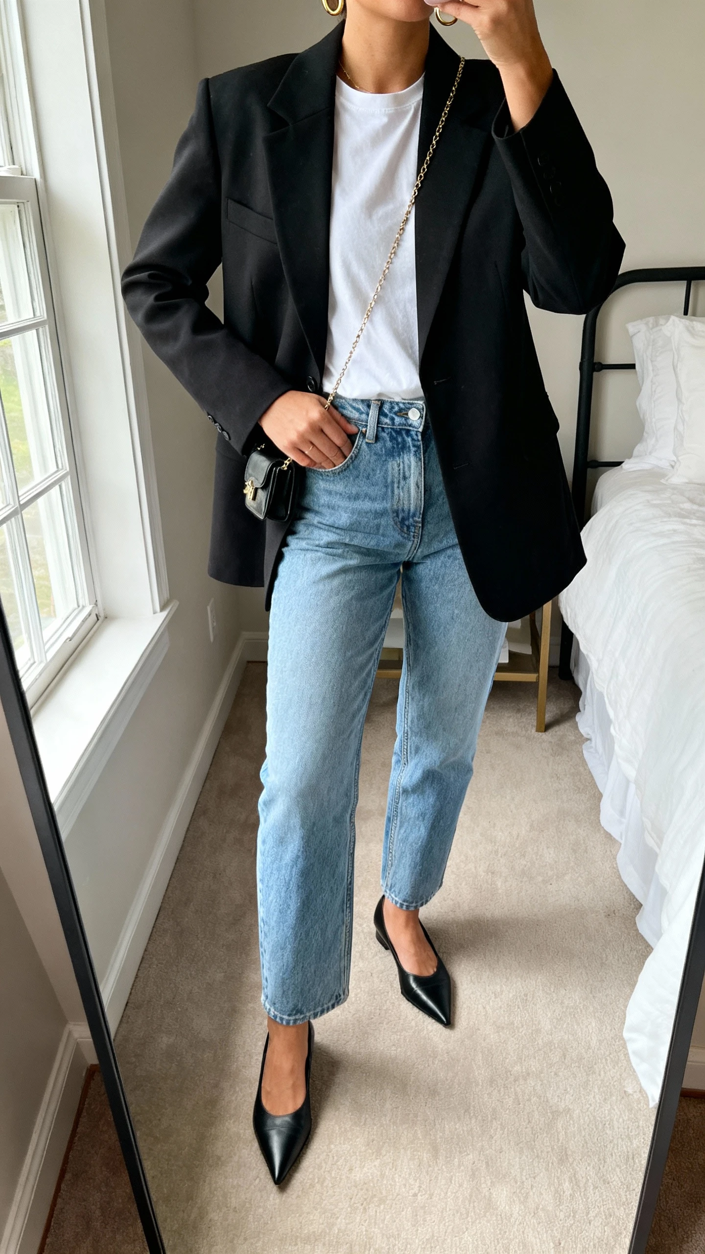 iPhone mirror selfie of a woman in a tailored black blazer over a white tee, light blue high-waisted straight-leg jeans, pointed-toe black flats, minimal gold hoops and thin chain, small crossbody in hand, face not visible, cropped at neck, bedroom mirror, soft window light, natural iPhone photo quality.