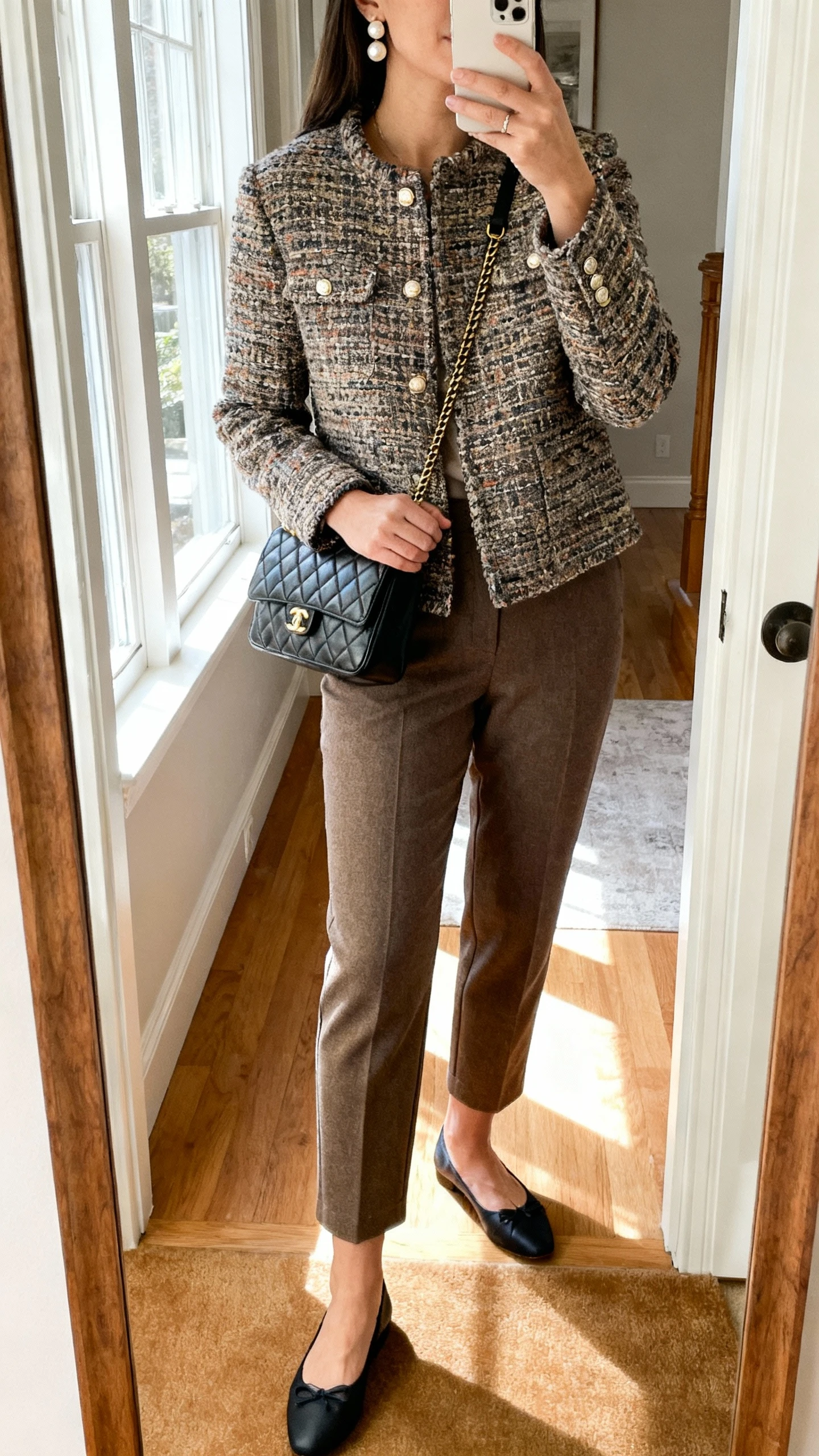 iPhone mirror selfie of a woman in a tweed jacket paired with tailored ankle pants, ballet flats, quilted shoulder bag, pearl earrings, face not visible, cropped at shoulders, entryway mirror, morning window light, natural iPhone photo quality.