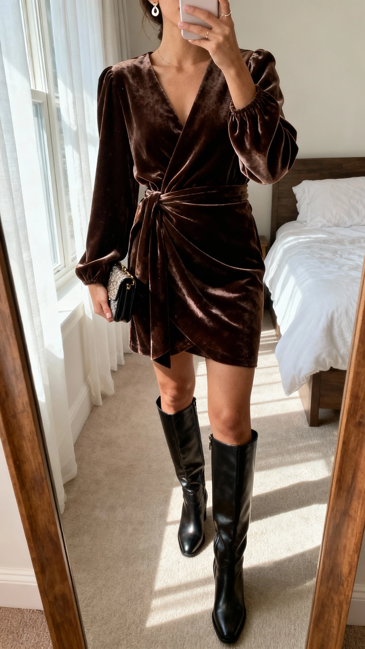 iPhone mirror selfie of a woman in a velvet wrap dress with sleek knee-high boots, dainty earrings and a small clutch, face not visible, cropped at neck, bedroom mirror with soft window light, natural iPhone photo quality.