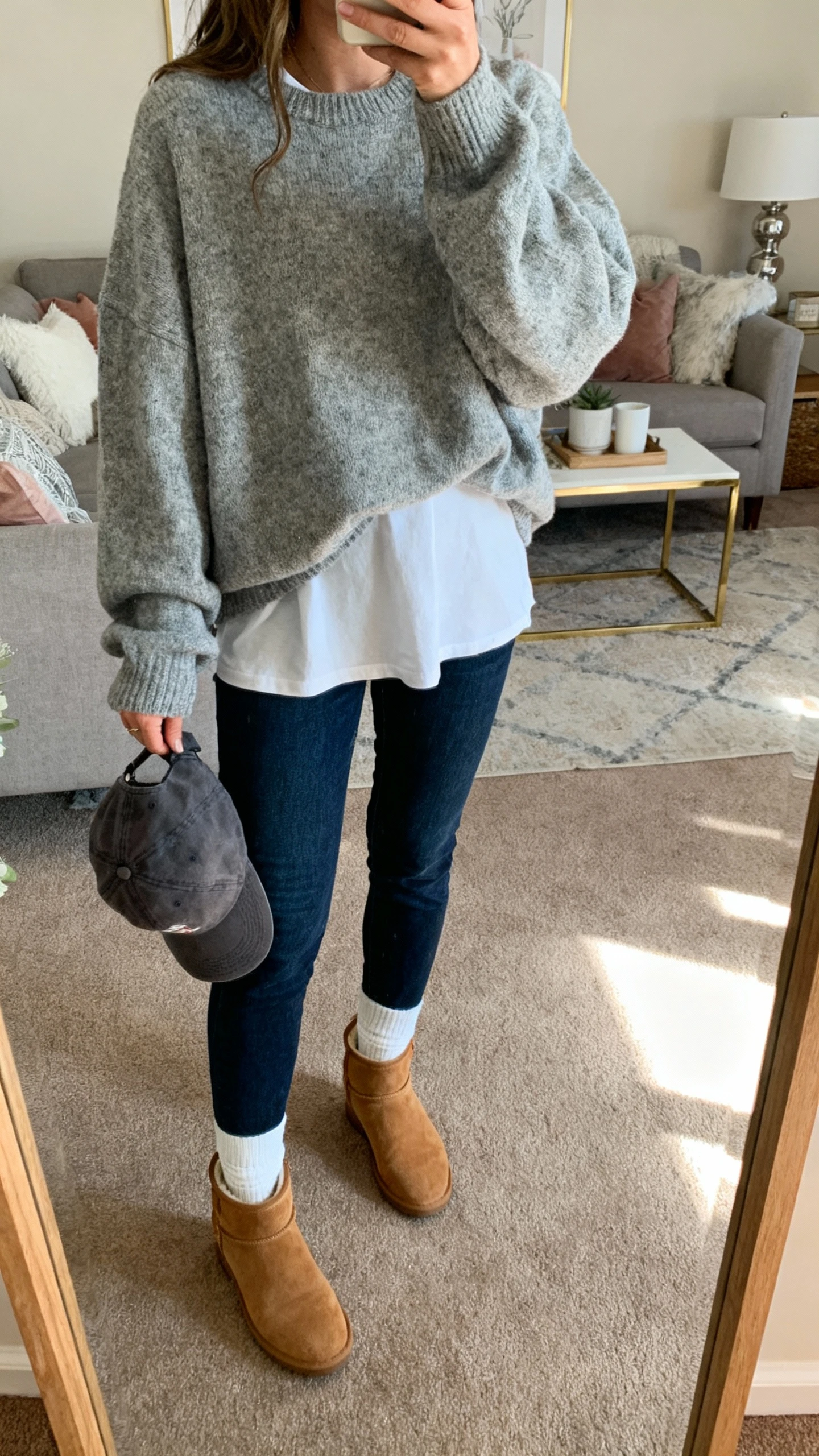 iPhone mirror selfie of a woman in an oversized heather gray sweater layered over a white tee, dark blue jeans, white crew socks, tan suede ankle boots, baseball cap in hand, face not visible, living room mirror with cozy weekend vibe, soft afternoon light, natural iPhone photo quality.