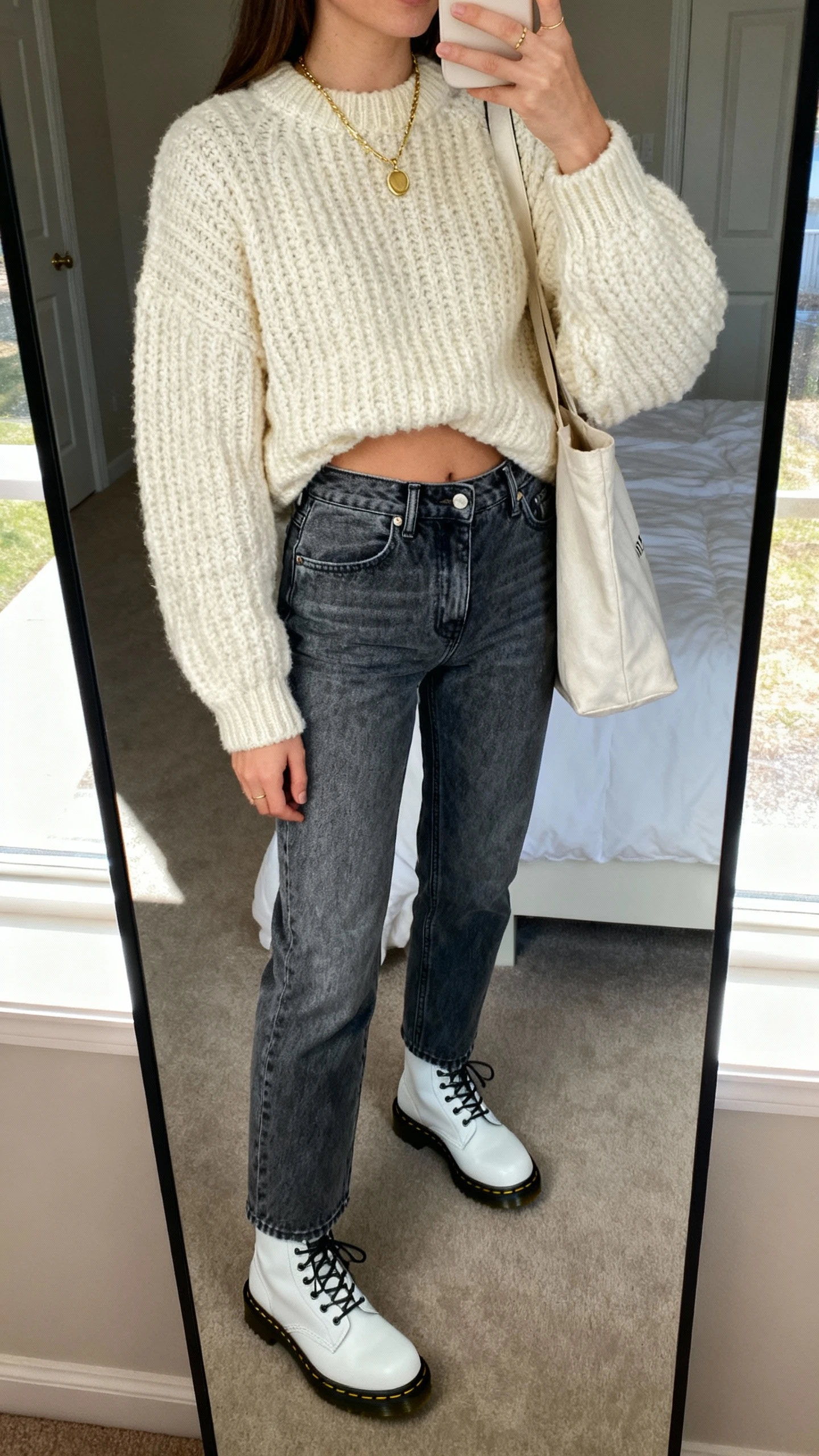 iPhone mirror selfie of a woman in cozy knit and denim: chunky cream sweater half-tucked into high-rise medium-wash jeans, white Doc Martens, delicate gold necklace, tote slung on shoulder; face not visible, bedroom full-length mirror, soft afternoon window light, casual iPhone photo.