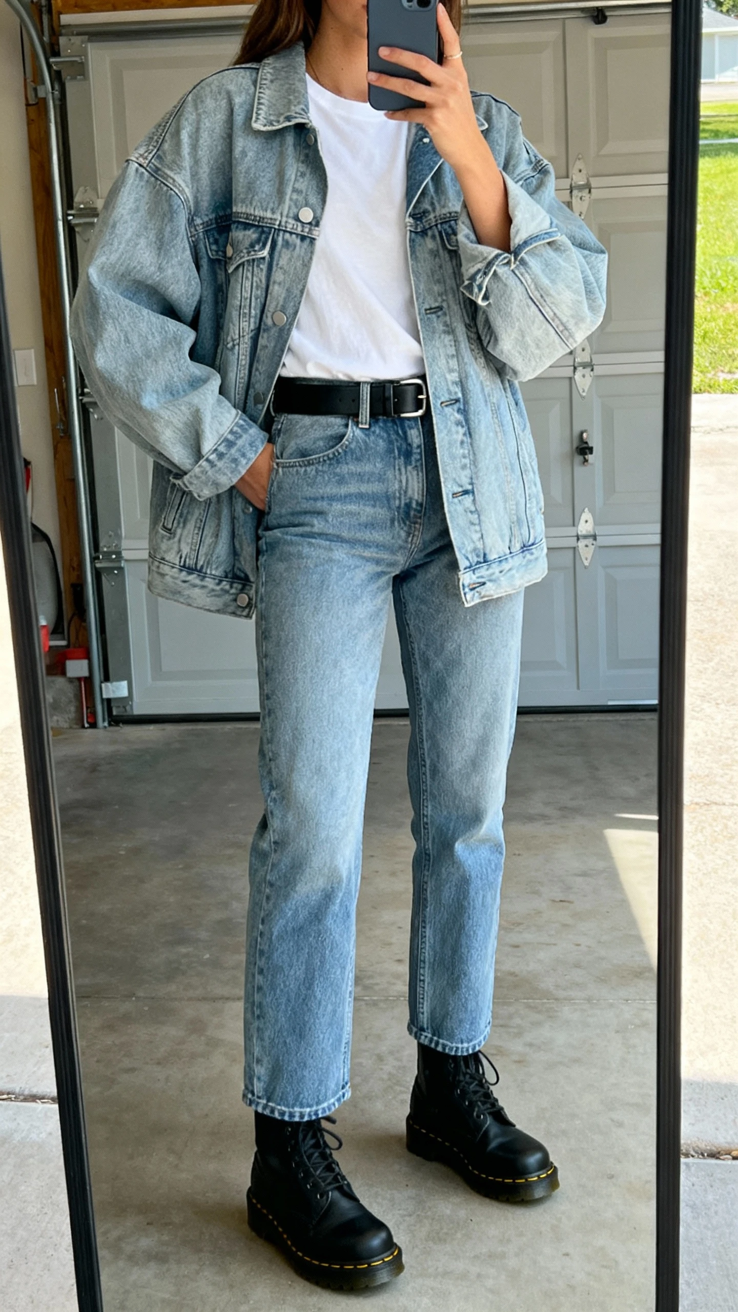 iPhone mirror selfie of a woman in denim-on-denim: light-wash oversized jean jacket with matching straight-leg jeans, white tee, black Doc Martens, black belt, face not visible, garage mirror, natural daylight, natural iPhone quality.