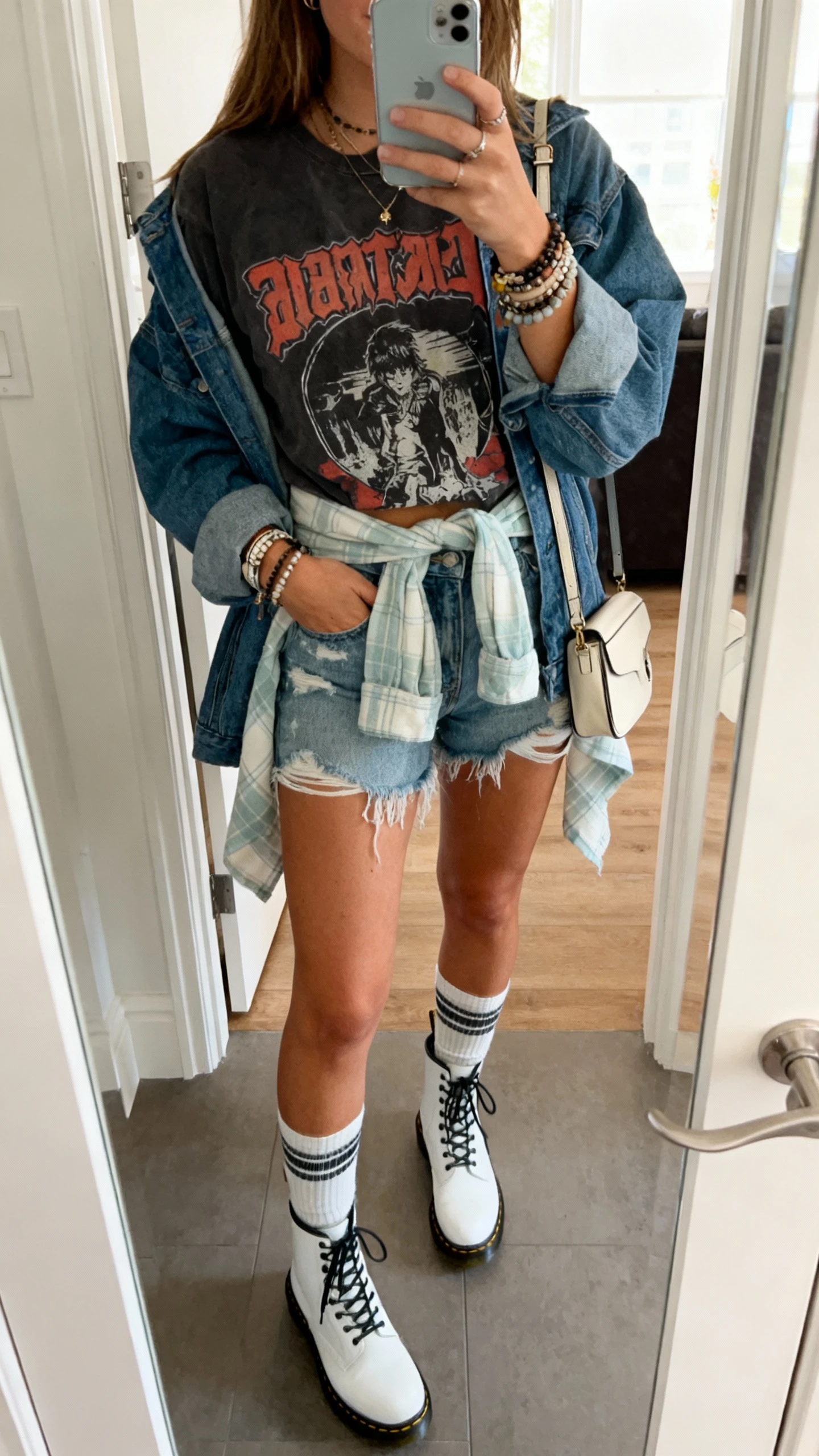 iPhone mirror selfie of a woman in festival-ready layers: distressed denim cutoffs, vintage band tee, light flannel tied at waist, oversized denim jacket, white Doc Martens with tall socks, layered bracelets and mini crossbody; face not visible, entryway mirror, natural morning light, casual iPhone vibe.