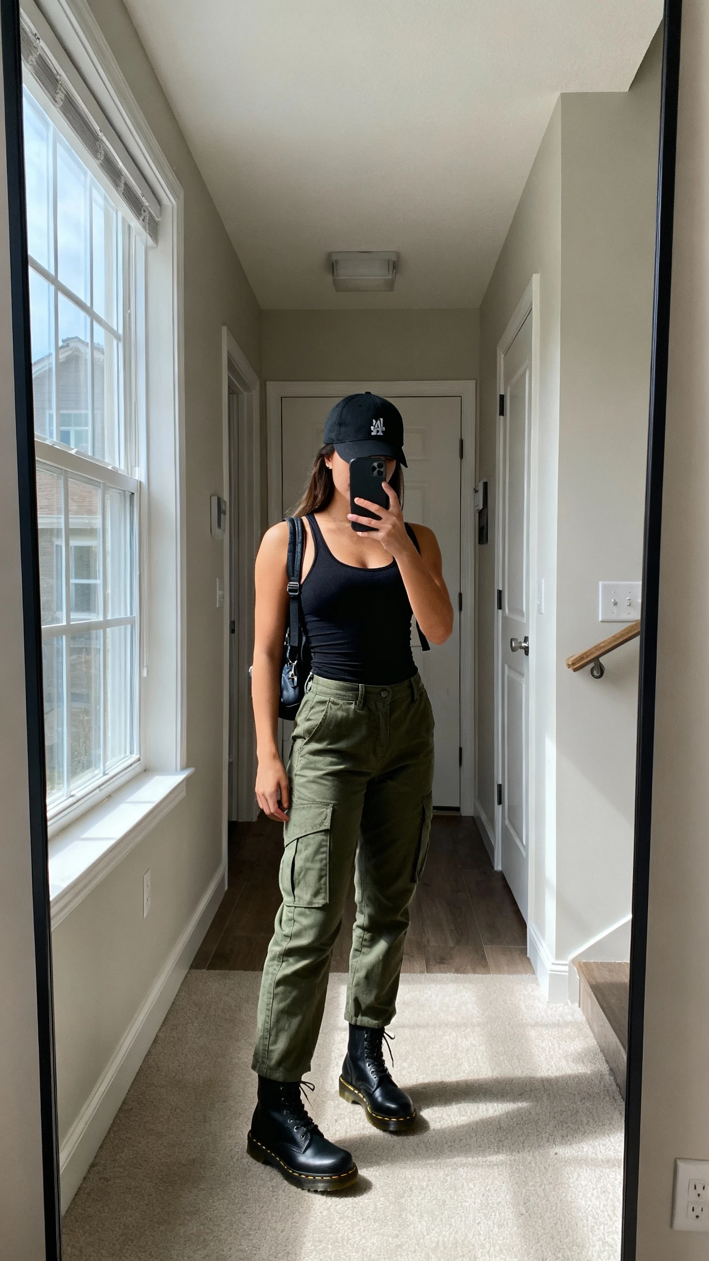 iPhone mirror selfie of a woman in olive cargo pants, black fitted tank, black Doc Martens, black baseball cap and small backpack, face not visible, apartment entryway mirror, daylight from nearby window, natural iPhone photo quality.