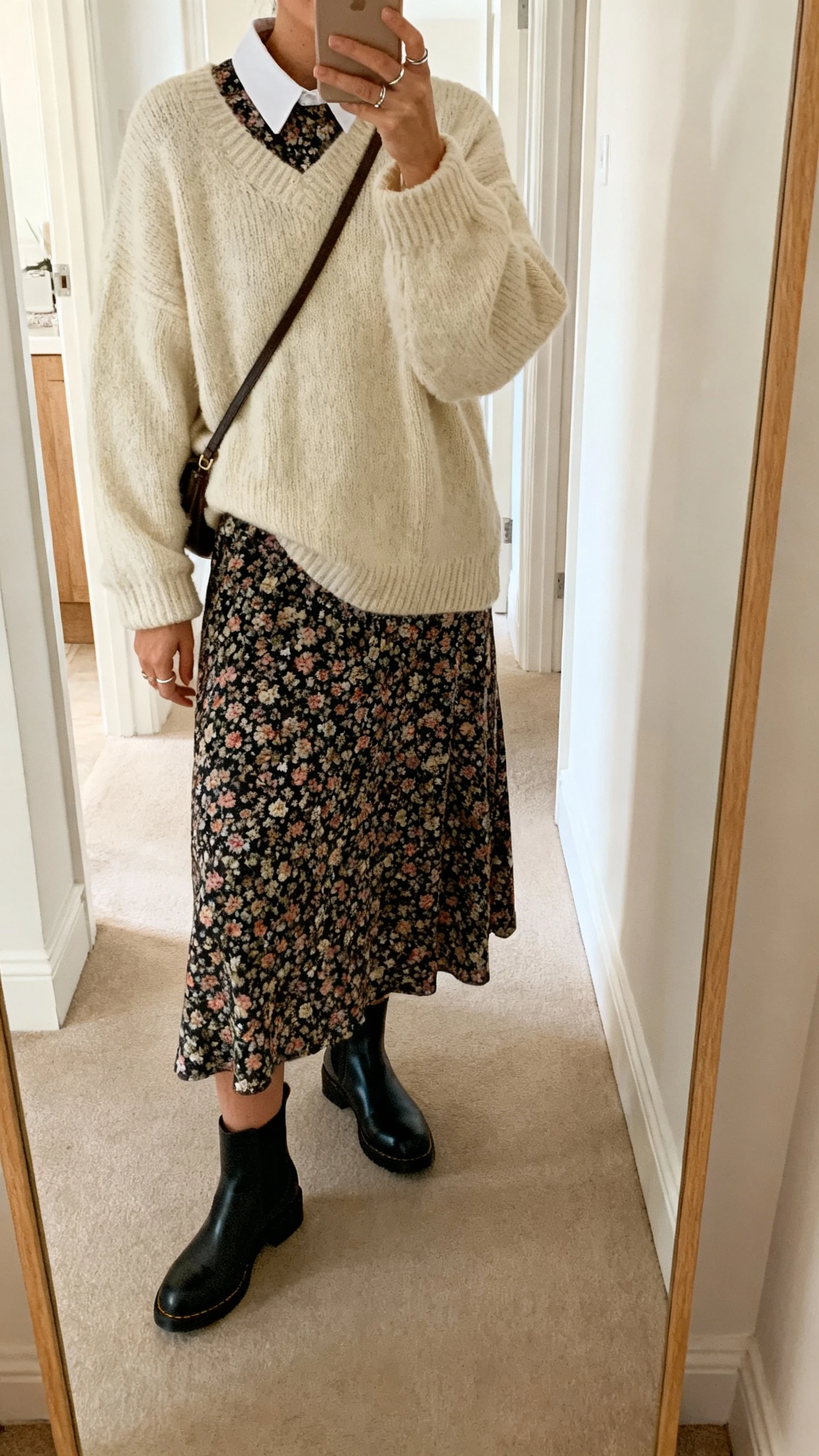 iPhone mirror selfie of a woman layering a chunky cream sweater over a floral midi dress (collar peeking), black Chelsea boots, crossbody bag, simple rings, face not visible, hallway full-length mirror, soft morning light, natural iPhone photo.
