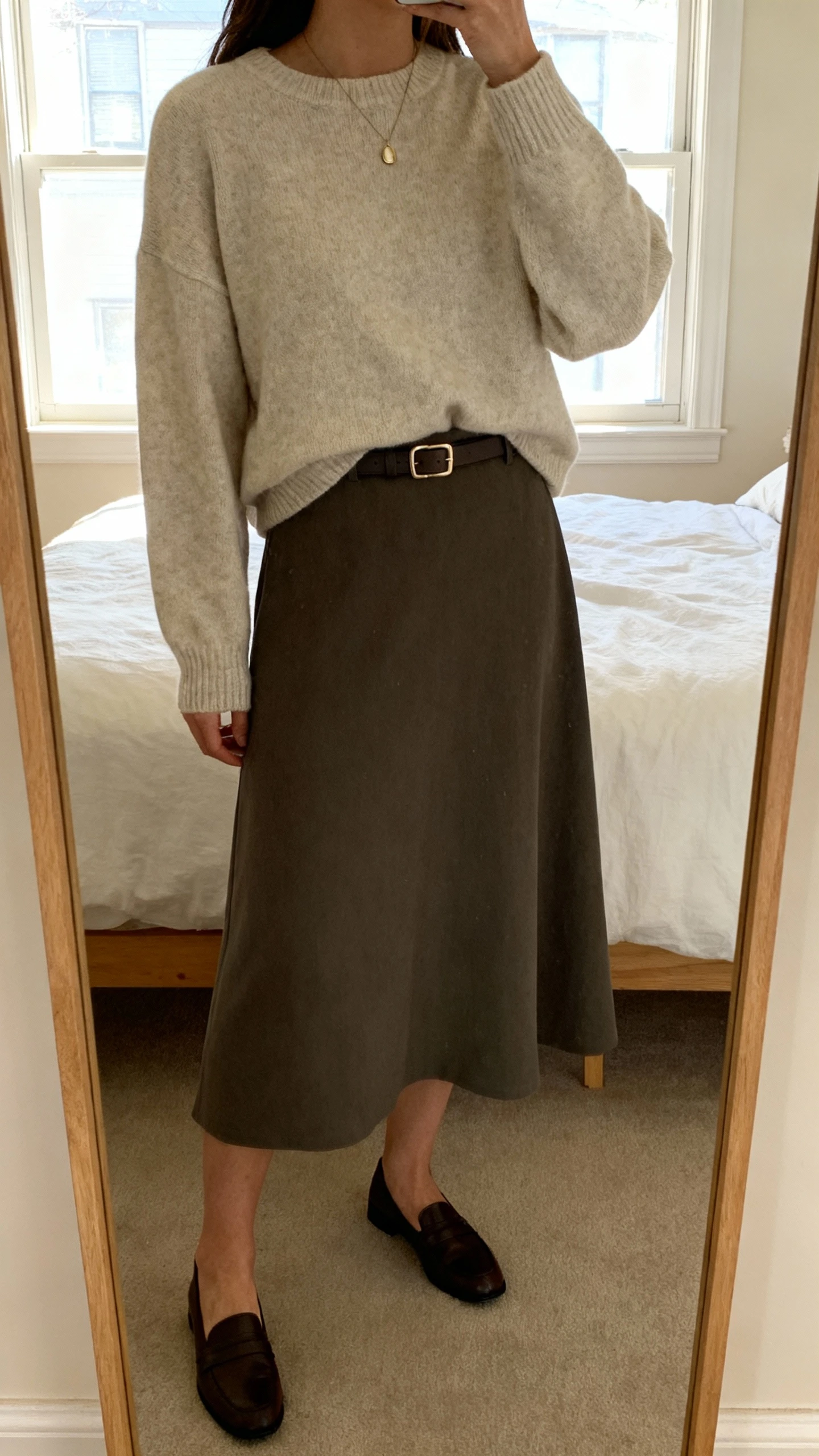iPhone mirror selfie of a woman layering a crewneck sweater over a midi dress, paired with Mary Janes or classic flats, slim belt at waist, simple pendant, face not visible, cropped at neck, bedroom mirror, morning window light, natural iPhone photo.