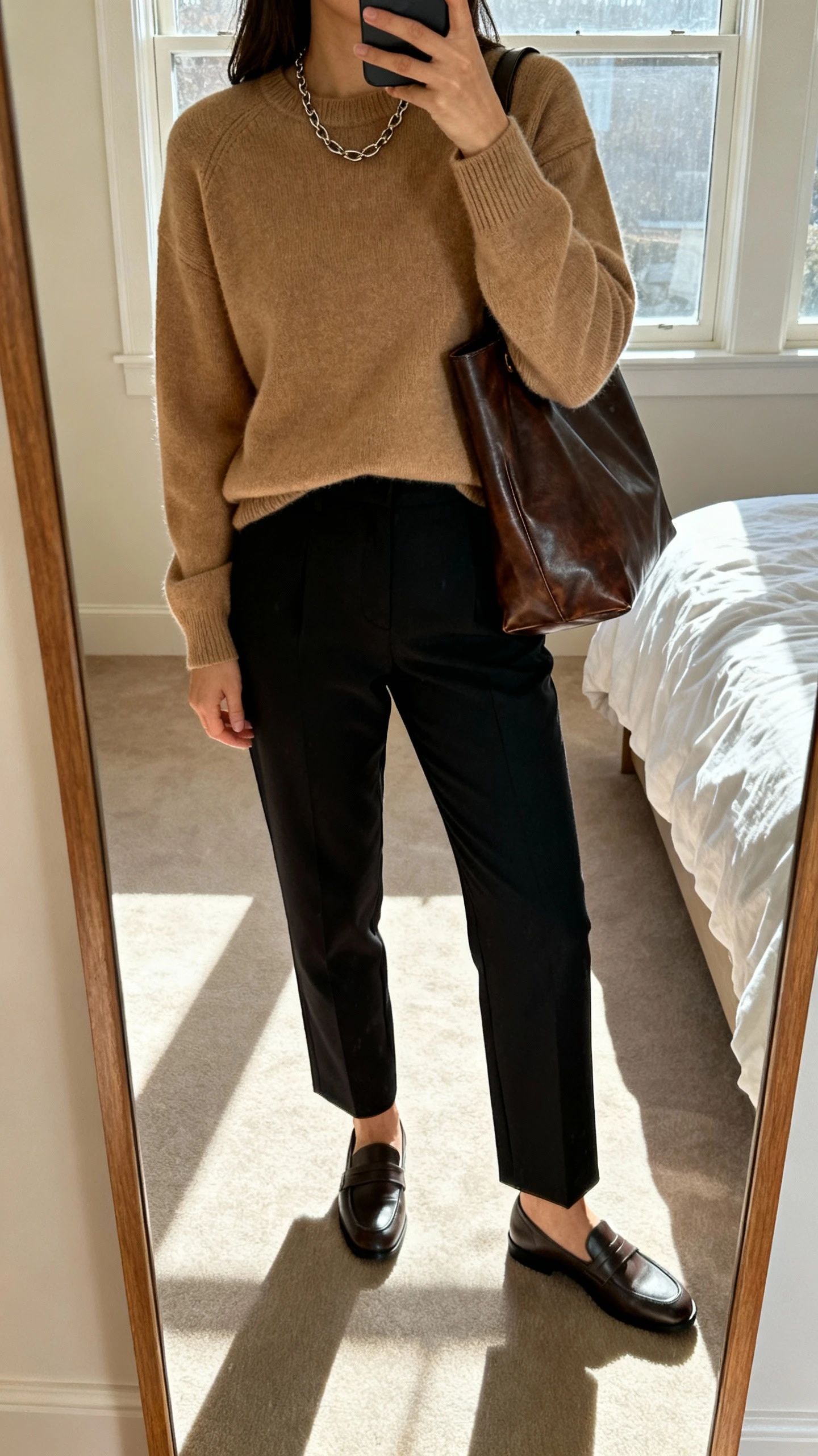 iPhone mirror selfie of a woman wearing a cashmere crewneck and trousers: camel cashmere crewneck, high-waist black tailored trousers, loafers, subtle chain necklace, leather tote, face not visible, cropped at shoulders, bedroom full-length mirror, morning window light, natural iPhone photo.