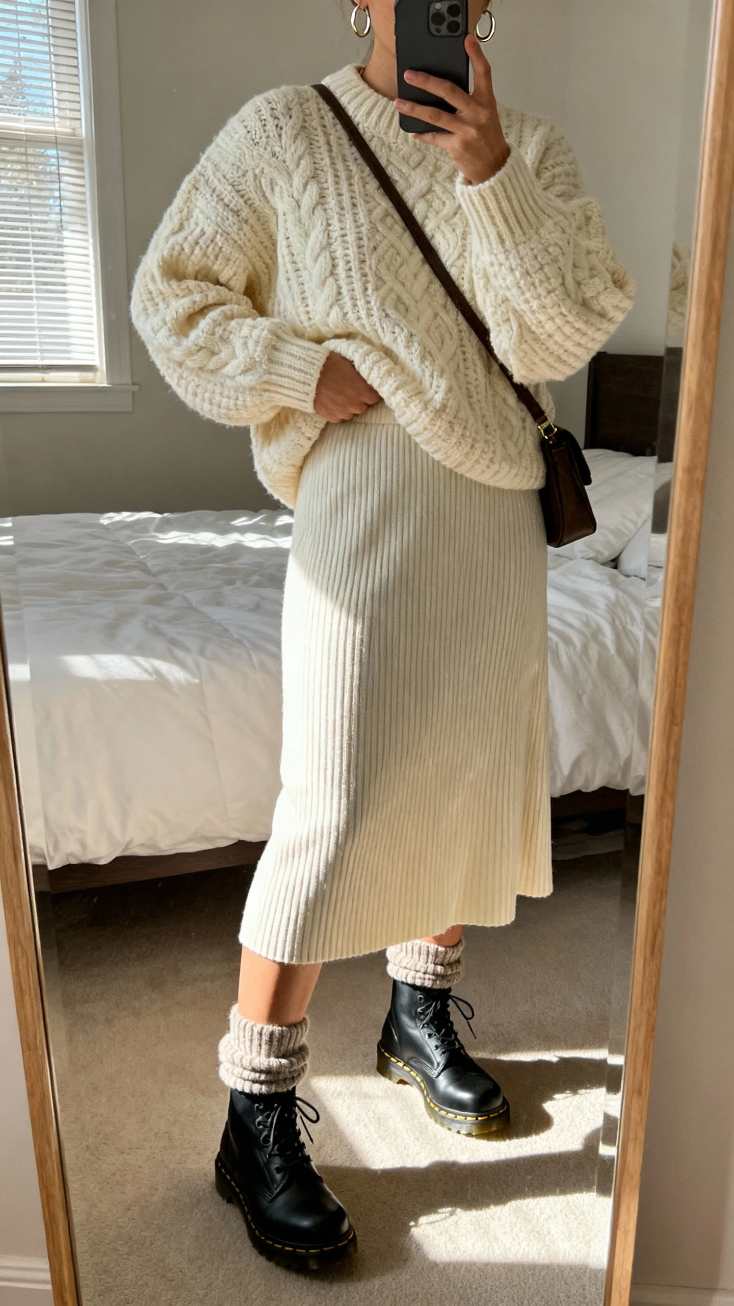 iPhone mirror selfie of a woman wearing a chunky cream knit sweater tucked into a ribbed midi skirt with black Doc Martens and slouchy wool socks, simple hoop earrings and a crossbody bag, face not visible, cropped at neck, bedroom mirror with morning window light, natural iPhone photo quality.