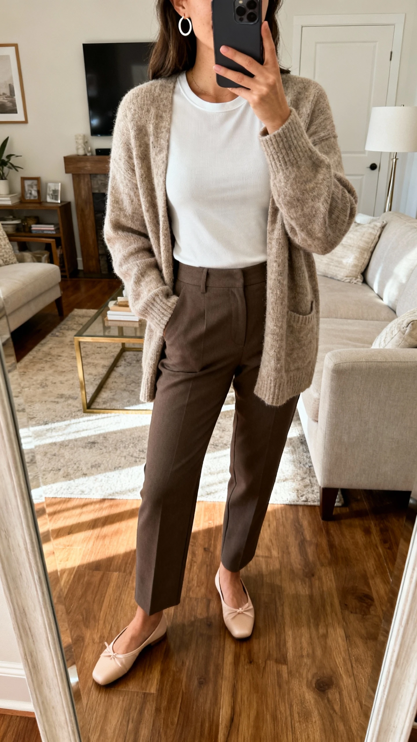 iPhone mirror selfie of a woman wearing a cozy cardigan over a fitted tee, tailored trousers, ballet flats, simple hoop earrings, face not visible, living room mirror with warm indoor light, natural iPhone photo quality.