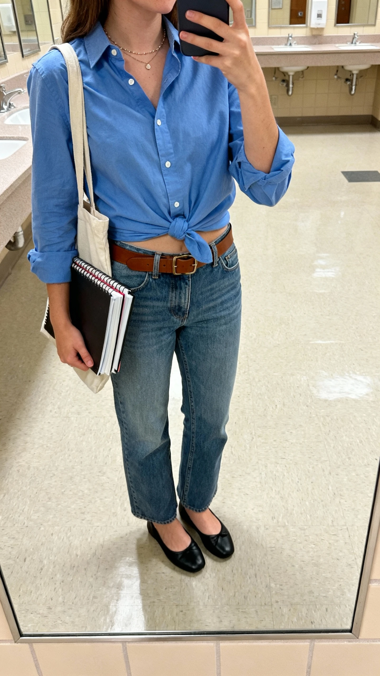 iPhone mirror selfie of a woman wearing a crisp blue button-down half-tucked into relaxed straight jeans, brown belt, black ballet flats, dainty necklace, tote with notebooks, face not visible, campus bathroom mirror, soft indoor lighting, iPhone photo quality.