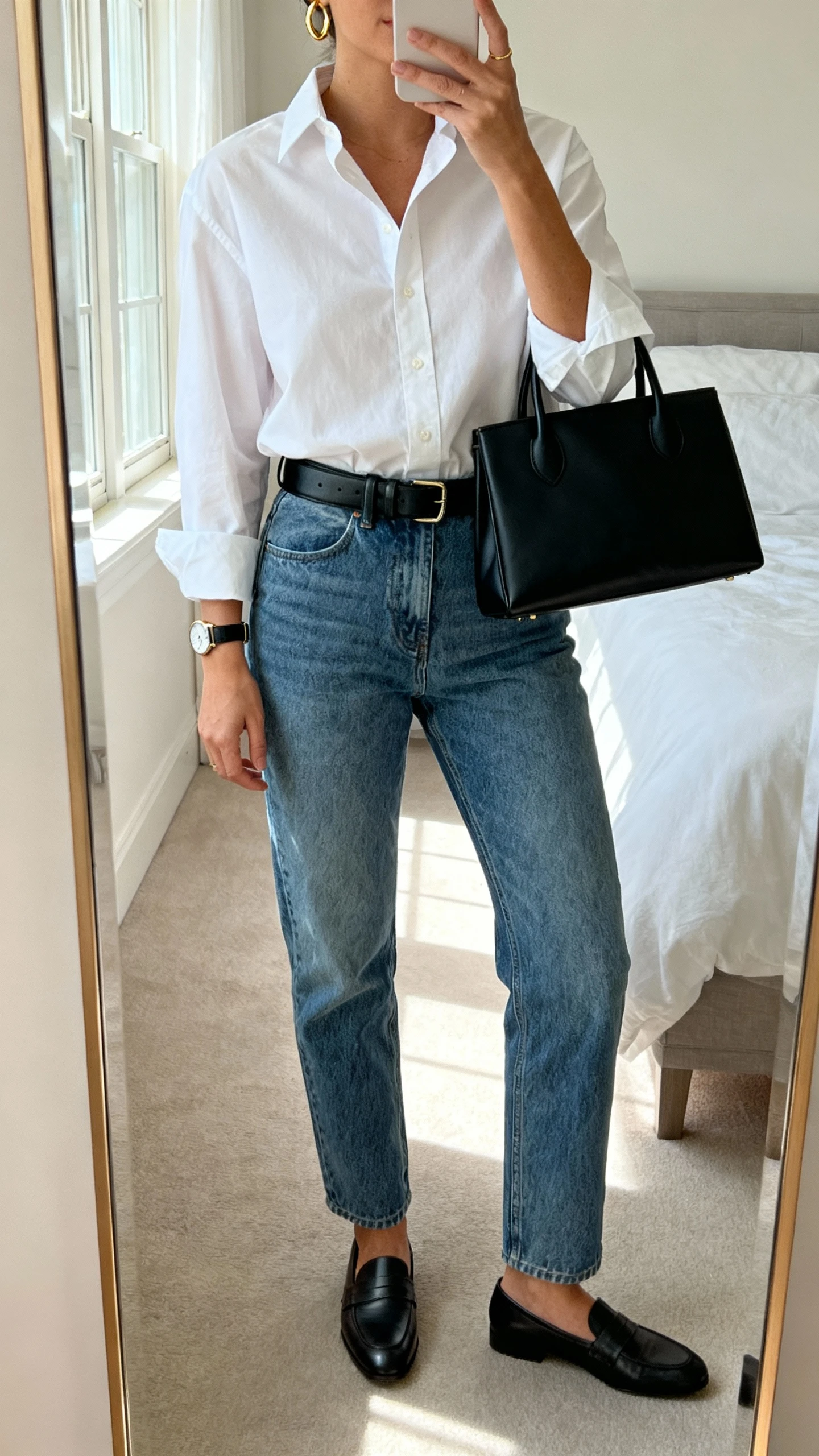 iPhone mirror selfie of a woman wearing a crisp white button-up shirt tucked into mid-wash blue jeans, black leather belt, loafers, simple gold hoops and watch, carrying a structured black tote, face not visible, cropped at neck, bedroom mirror, soft morning window light, natural iPhone photo quality.