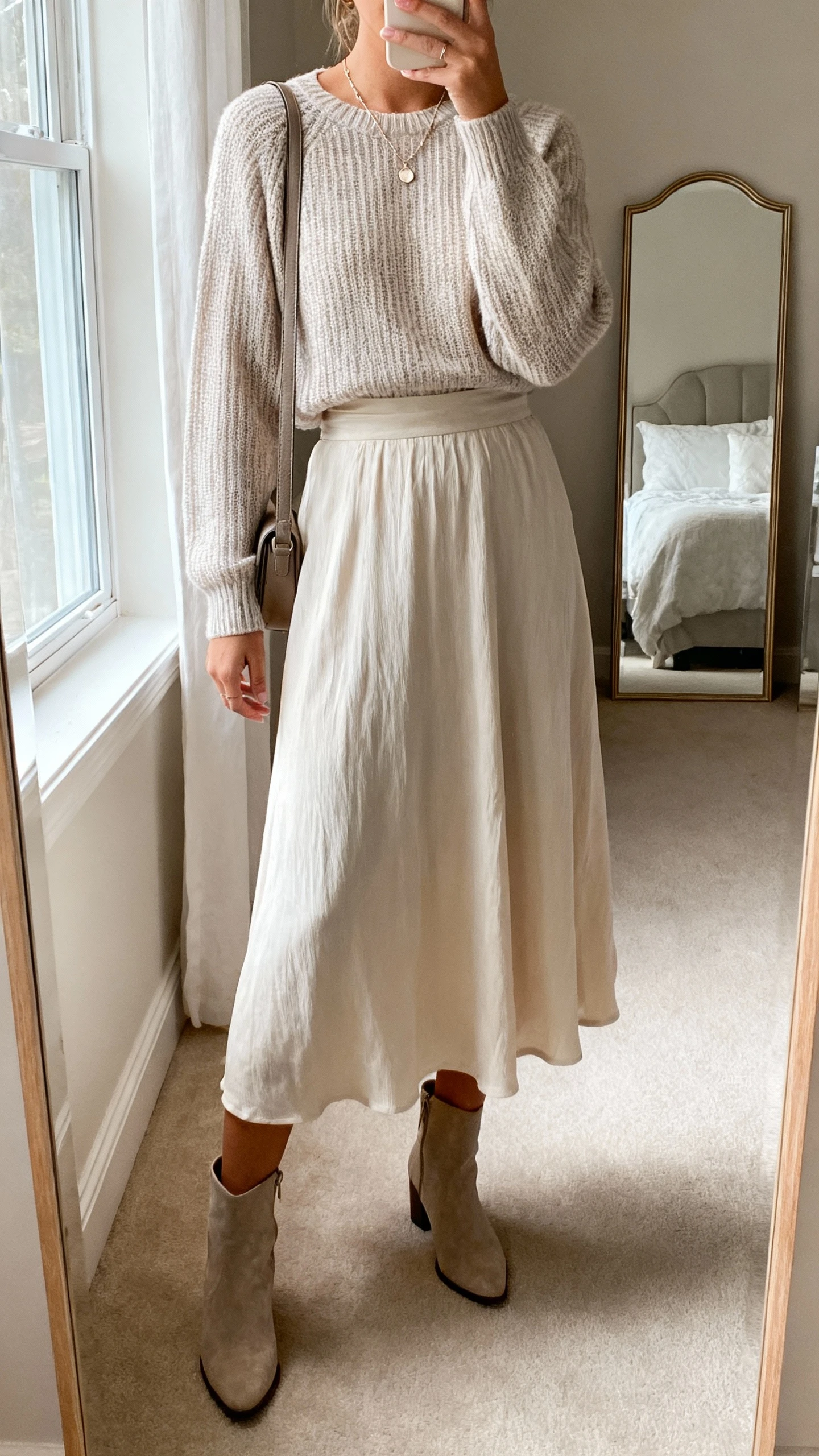 iPhone mirror selfie of a woman wearing a fine-knit sweater tucked into a flowy midi skirt, ankle boots, delicate necklace, and a crossbody bag, face not visible, bedroom mirror, soft natural window light, casual iPhone aesthetic