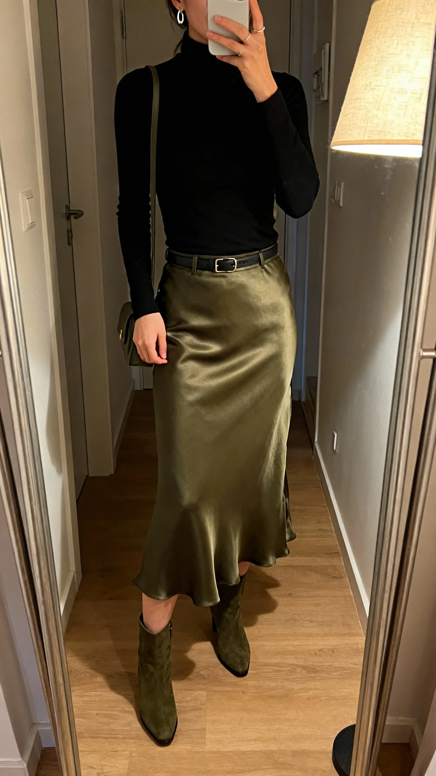 iPhone mirror selfie of a woman wearing a fitted black turtleneck tucked into a satin slip skirt in olive, ankle boots, delicate earrings, small shoulder bag, face not visible, apartment hallway mirror, evening lamp light, natural iPhone photo.