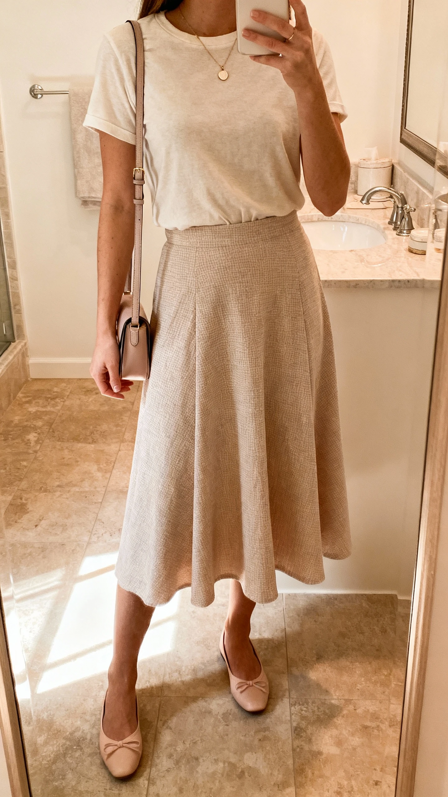 iPhone mirror selfie of a woman wearing a flirty A-line skirt with a tucked-in soft cotton tee, dainty pendant necklace, ballet flats, small shoulder bag; face not visible, bathroom mirror, warm morning light, natural iPhone photo quality