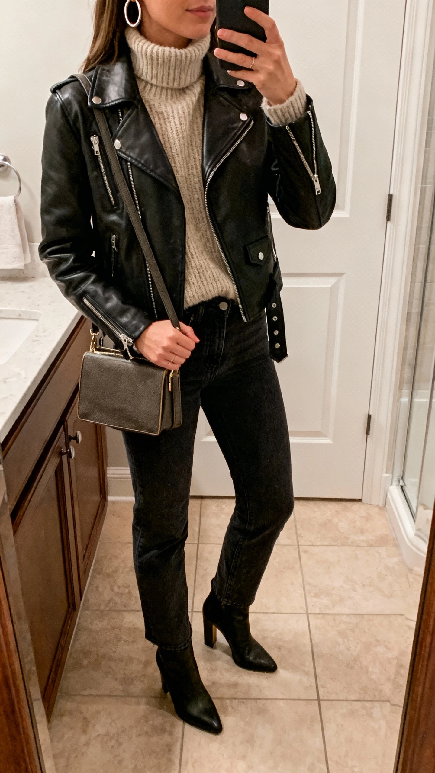 iPhone mirror selfie of a woman wearing a leather moment with cozy knits: black leather moto jacket, chunky oatmeal turtleneck, dark straight-leg jeans, heeled boots, hoop earrings, structured crossbody, face not visible, bathroom mirror with warm indoor lighting, natural iPhone aesthetic.