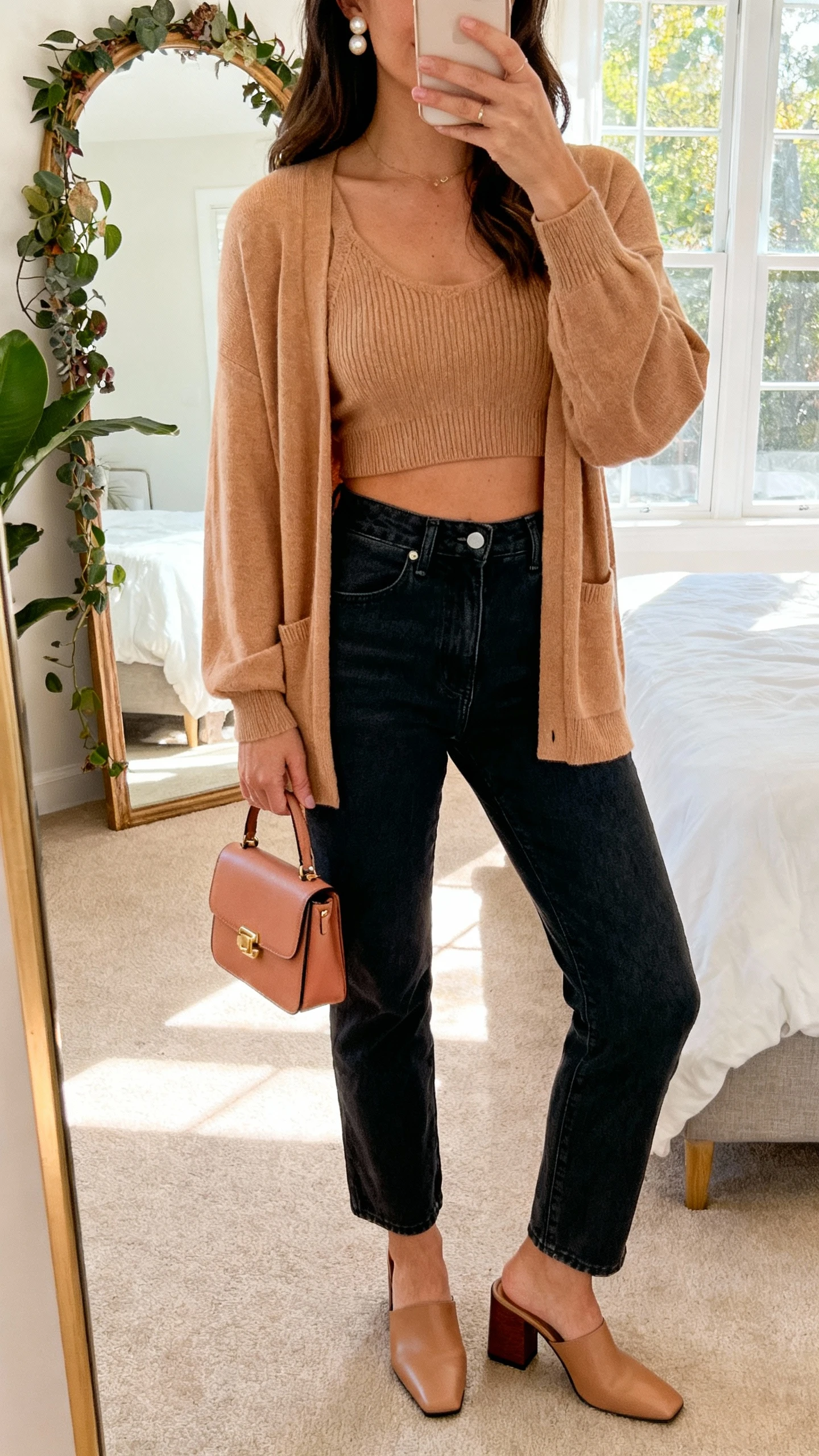iPhone mirror selfie of a woman wearing a matching cardigan set (cropped knit tank and cardigan) in soft tan, high-waisted dark jeans, block-heel mules, pearl studs, mini tote, face not visible, bedroom mirror with plants, afternoon window light, natural iPhone photo.