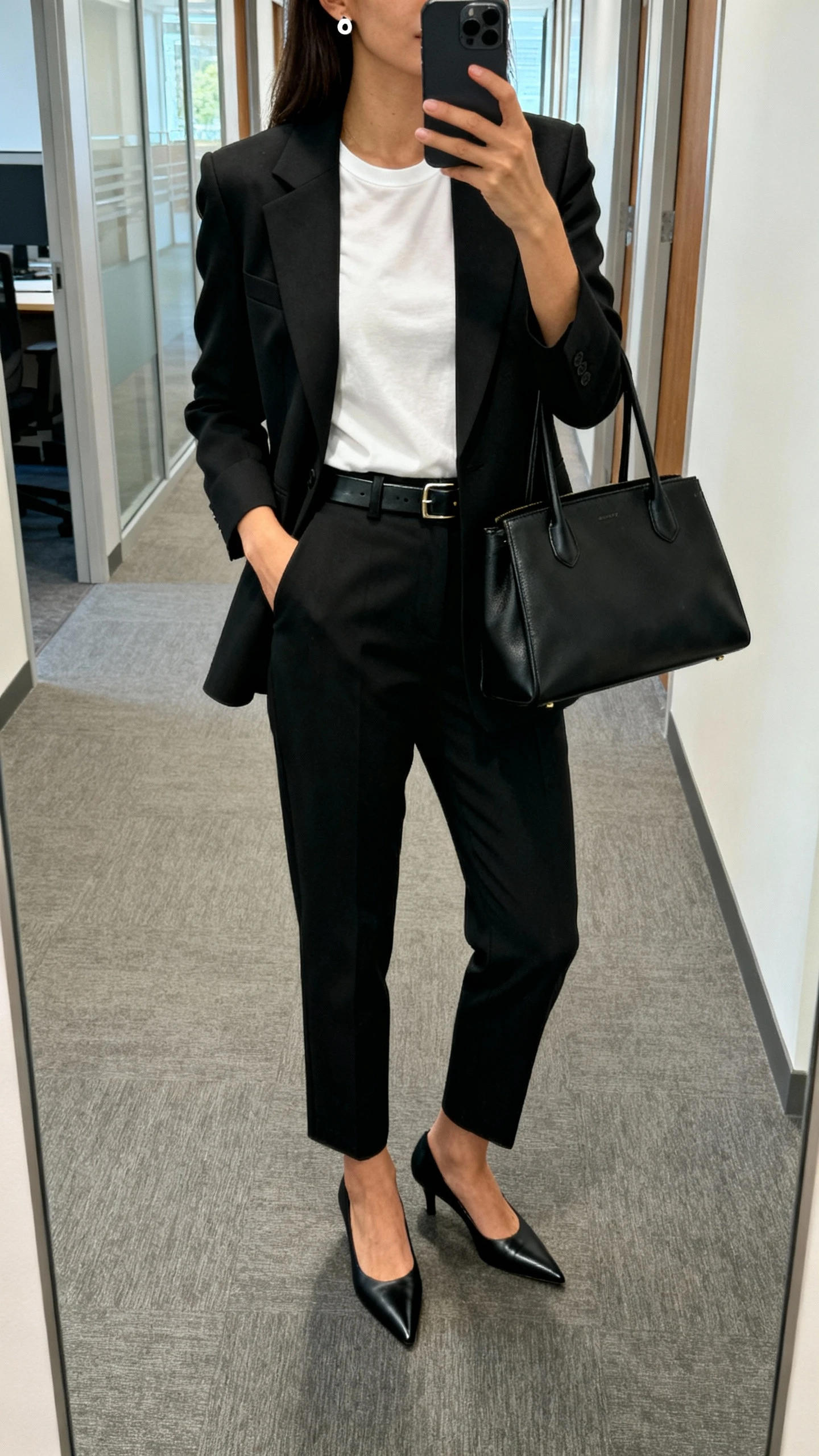 iPhone mirror selfie of a woman wearing a modern monochrome suit: tailored black blazer, matching straight-leg trousers, crisp white tee, black pointed pumps, slim leather belt, minimal stud earrings, structured black tote, face not visible, cropped at neck, office hallway mirror, natural morning light, natural iPhone photo quality.