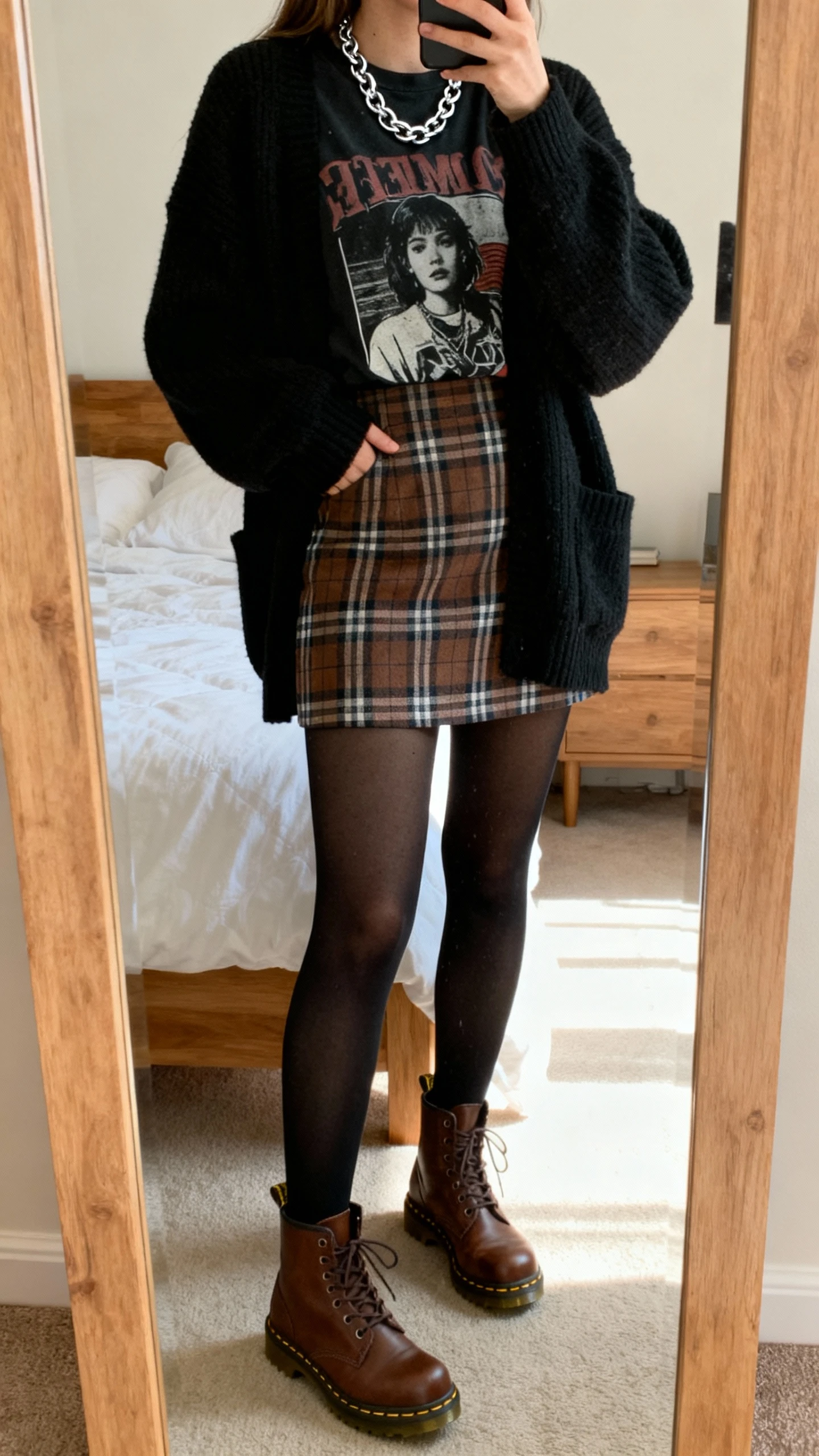 iPhone mirror selfie of a woman wearing a plaid mini skirt, black tights, brown Doc Martens, oversized black cardigan over a graphic tee, and silver chain necklace; face not visible, bedroom full-length mirror, afternoon natural light, casual iPhone quality.