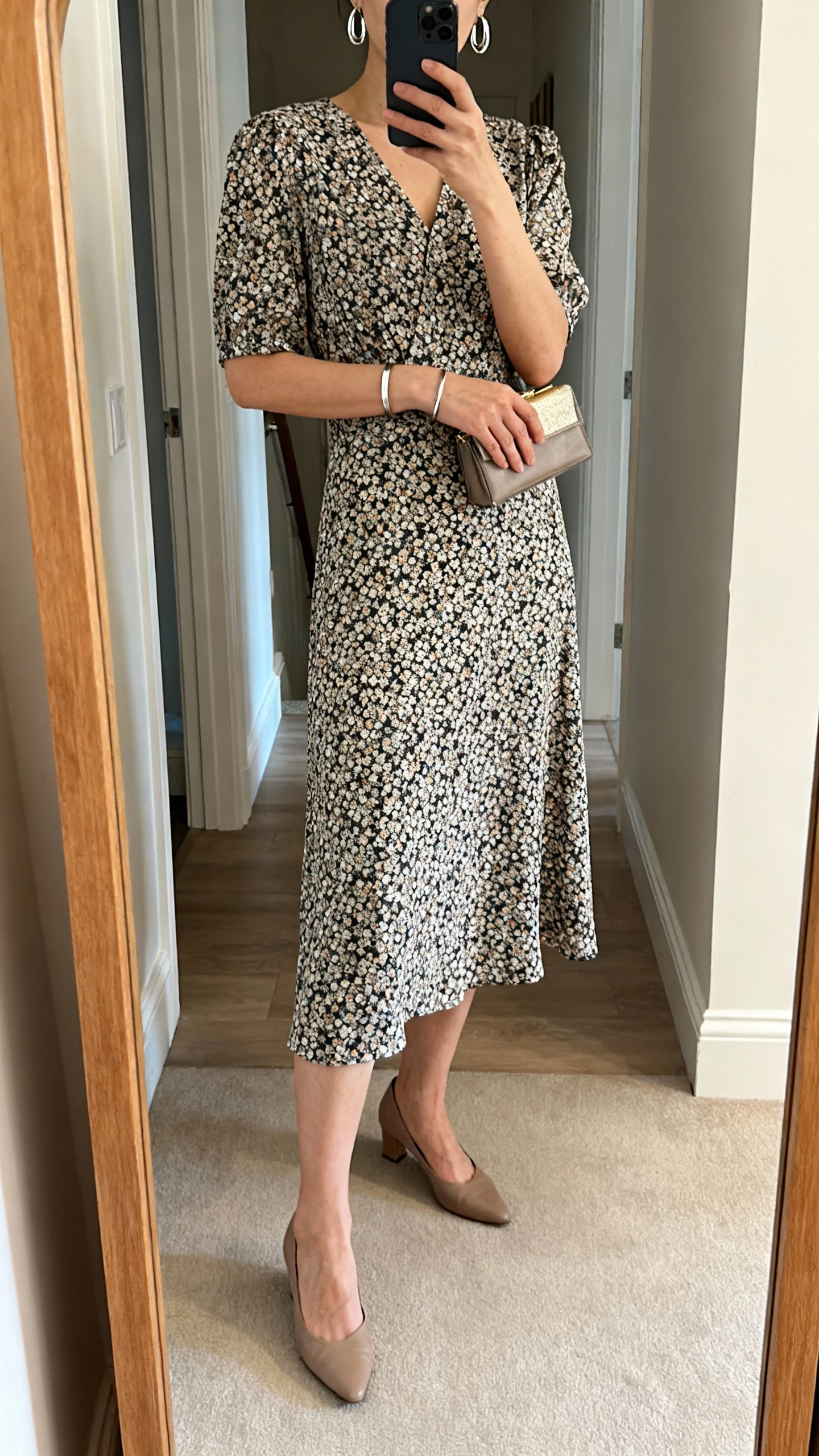 iPhone mirror selfie of a woman wearing a printed midi dress with low heels, subtle shine via metallic earrings and a slim bracelet, small clutch, face not visible, cropped at neck, entryway mirror, soft indoor lighting, natural iPhone photo quality.