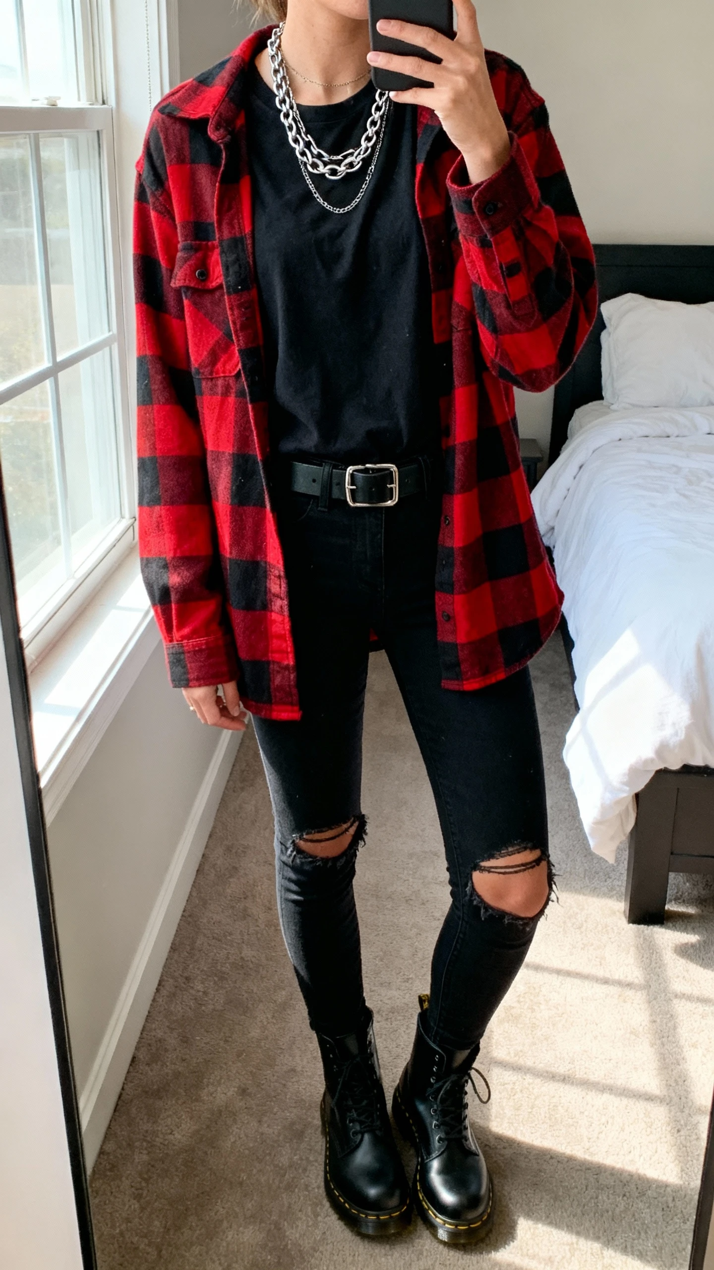 iPhone mirror selfie of a woman wearing a red-and-black flannel over a black tee, ripped black skinny jeans, black Doc Martens, layered silver chain necklace, black belt, face not visible, cropped at neck, bedroom mirror with window light, natural iPhone photo quality.