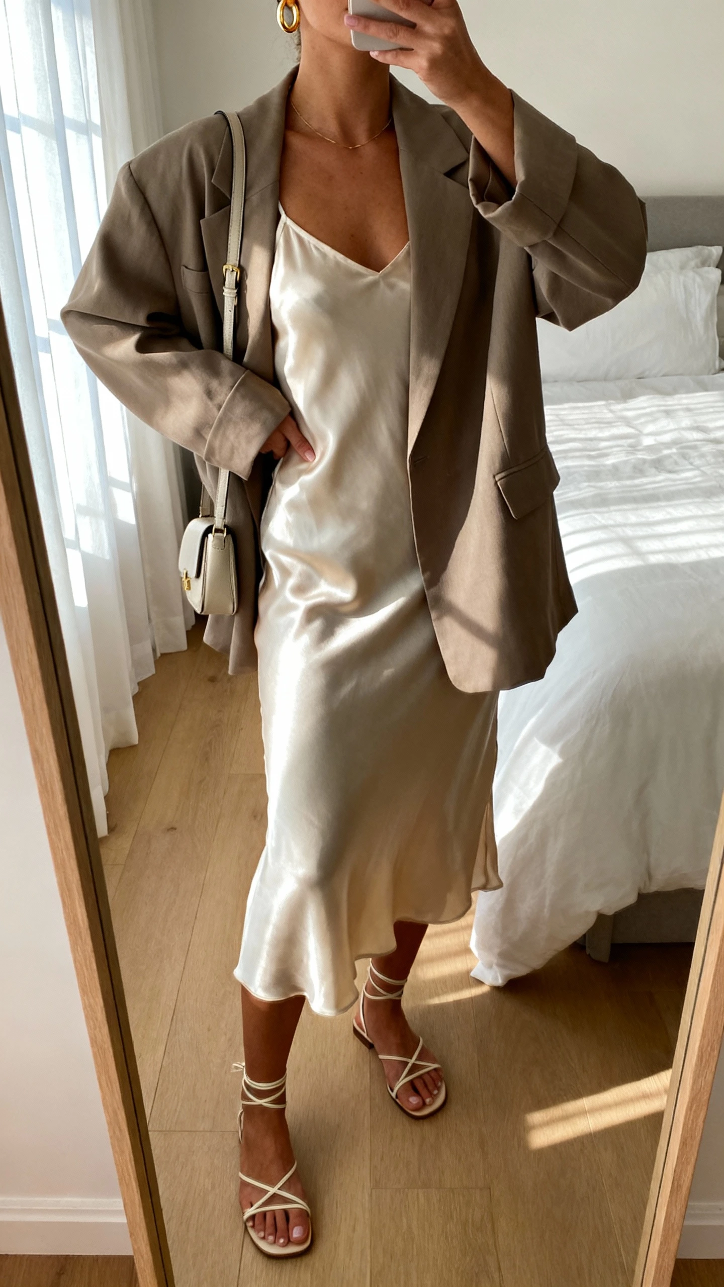 iPhone mirror selfie of a woman wearing a silky slip dress with a relaxed oversized blazer, minimalist strappy sandals, delicate gold hoops and a small shoulder bag, face not visible, cropped at neck, bedroom mirror with soft morning window light, natural iPhone photo quality.