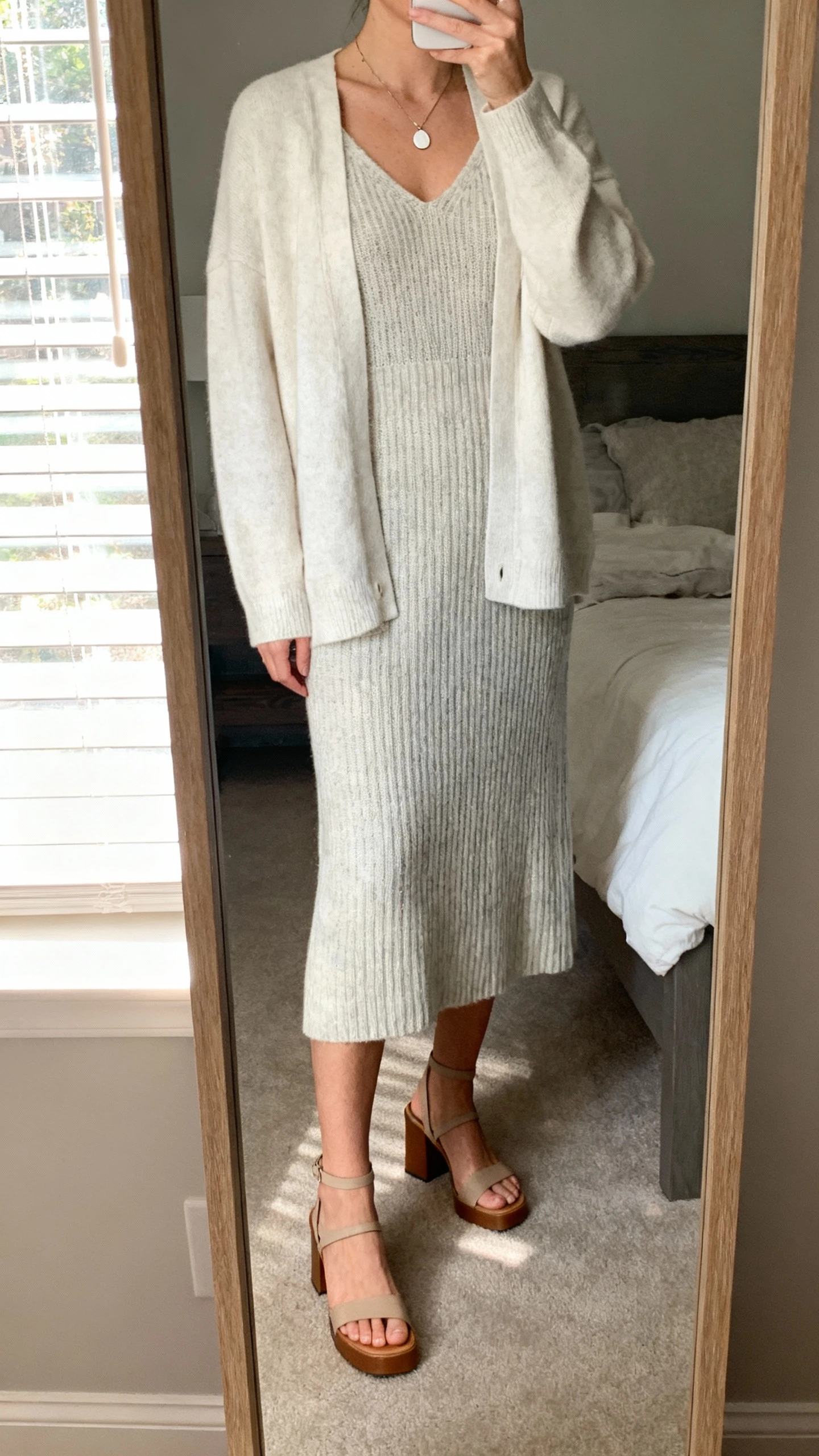 iPhone mirror selfie of a woman wearing a soft knit midi dress layered with a light cardigan and simple pendant necklace, low block-heel sandals, face not visible, cropped at neck, bedroom mirror with natural window light, natural iPhone photo quality.