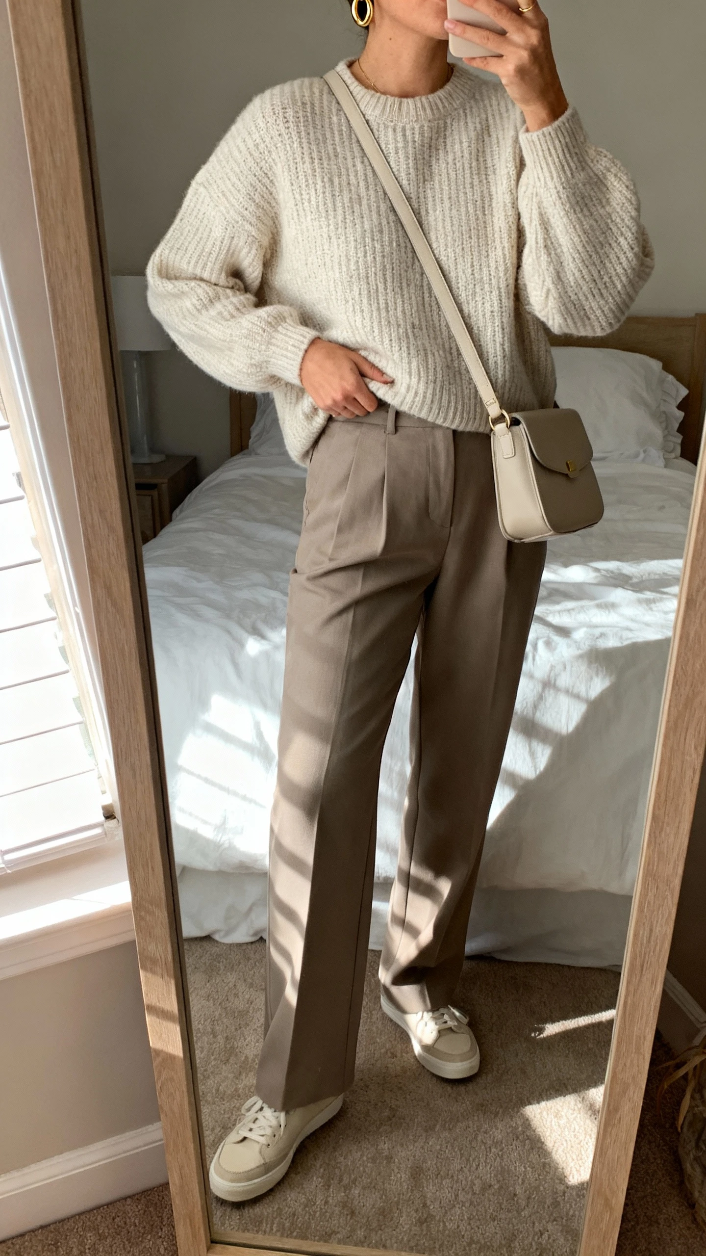 iPhone mirror selfie of a woman wearing a soft knit sweater tucked into tailored trousers with minimalist sneakers, delicate gold hoops, simple crossbody bag, face not visible, cropped at neck, bedroom mirror with morning window light, natural iPhone photo quality.