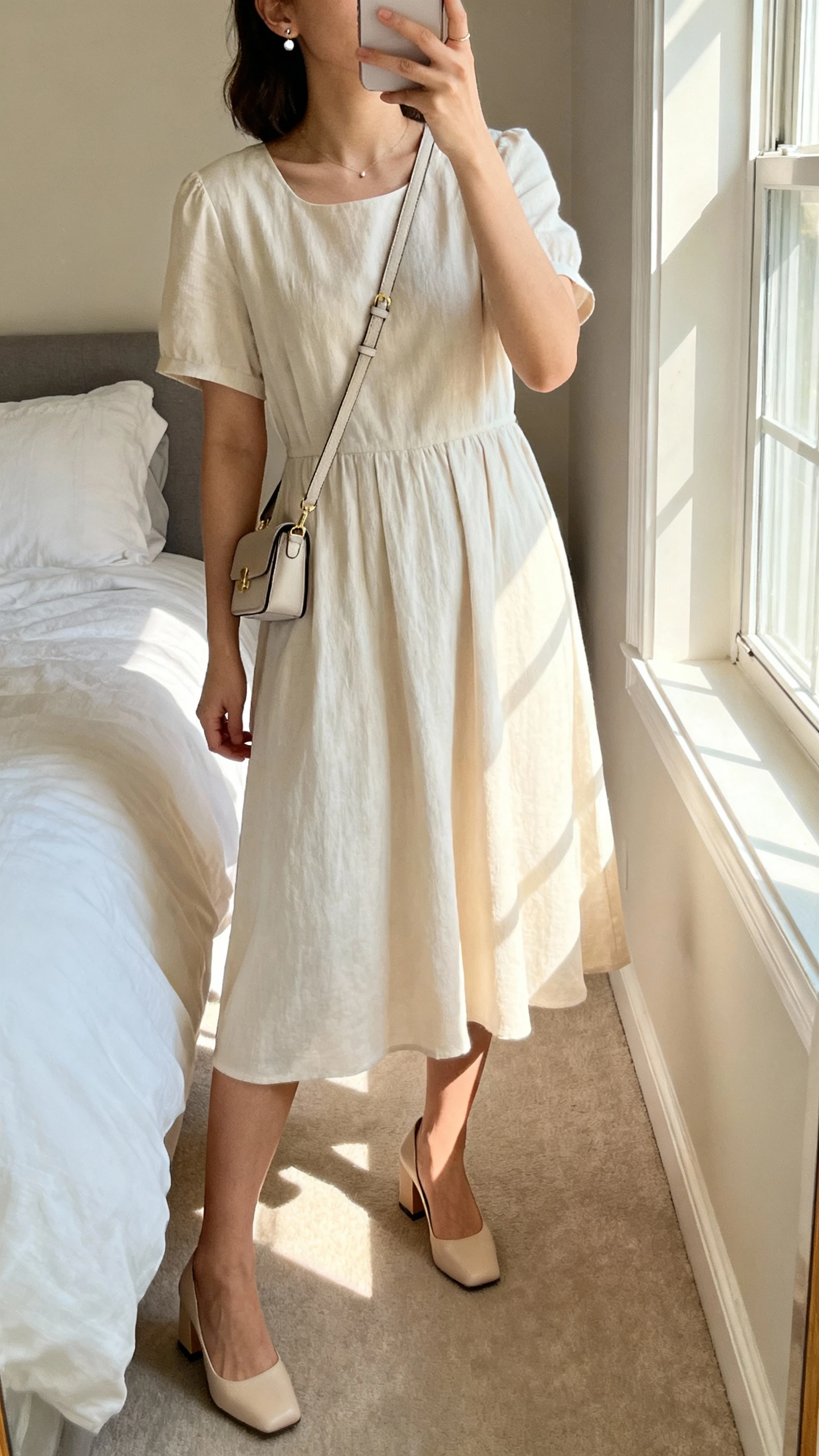iPhone mirror selfie of a woman wearing a soft midi dress with block heels, delicate stud earrings, small crossbody bag, face not visible, cropped at neck, bedroom mirror with morning window light, natural iPhone photo quality.