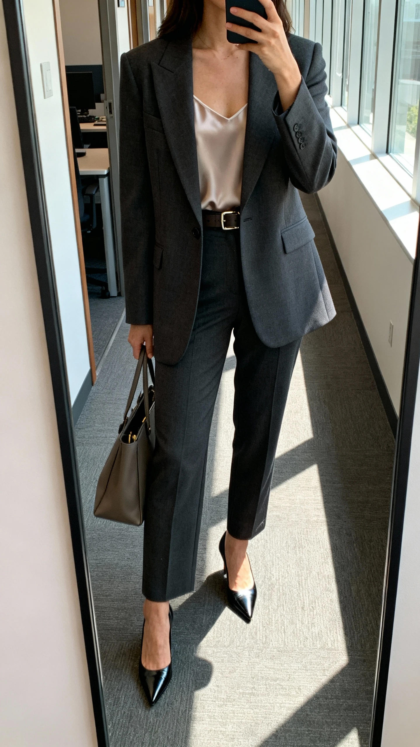 iPhone mirror selfie of a woman wearing a structured power blazer, tailored trousers, pointed-toe pumps, simple silk camisole, slim belt, and a tote, face not visible, cropped at neck, office hallway mirror, morning window light, natural iPhone photo quality