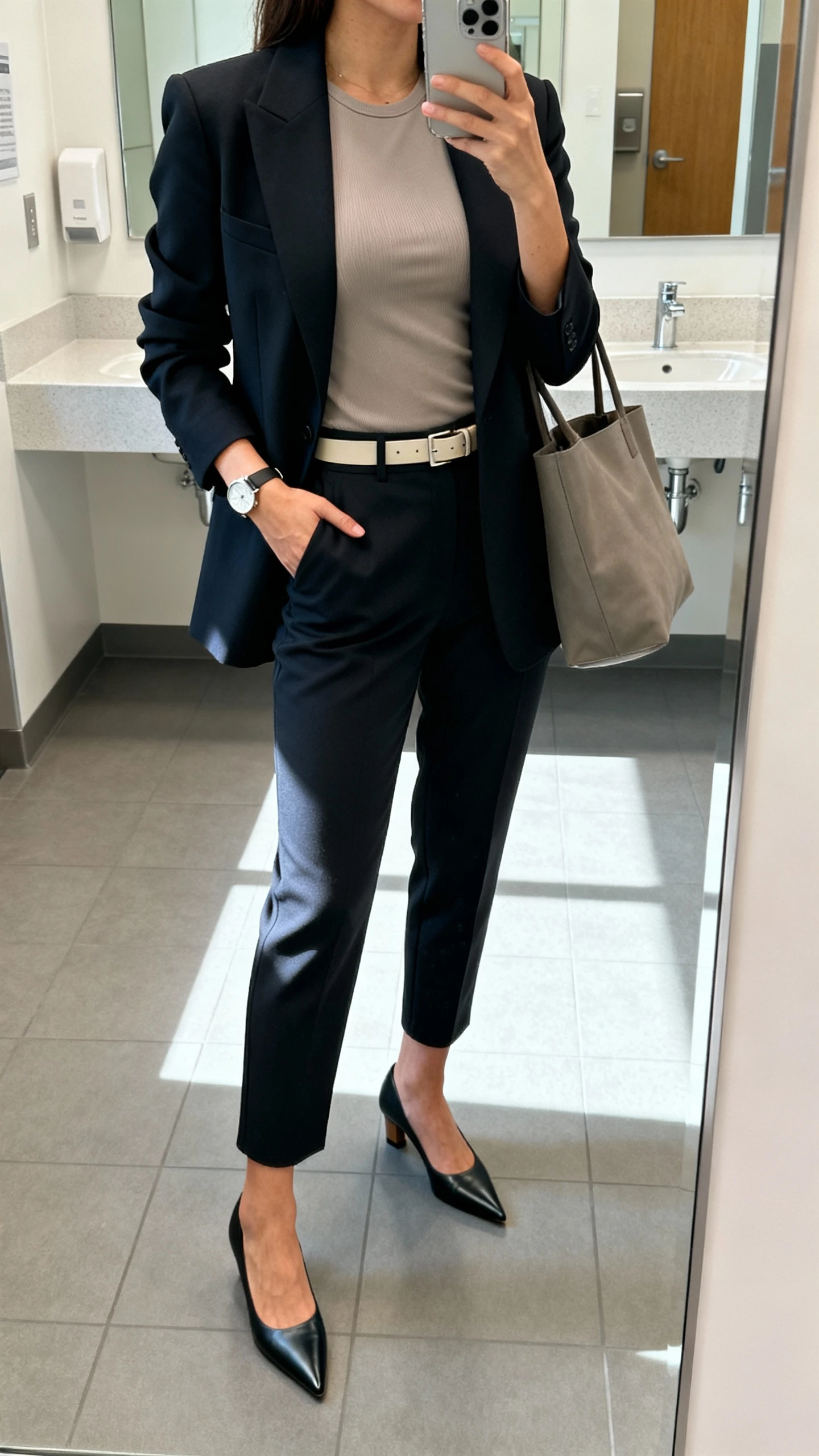 iPhone mirror selfie of a woman wearing a structured power blazer over a fitted shell top, tapered trousers, pointed-toe pumps, slim belt, minimalist watch, and tote bag, face not visible, cropped at neck, office restroom mirror, bright morning light, natural iPhone photo quality.