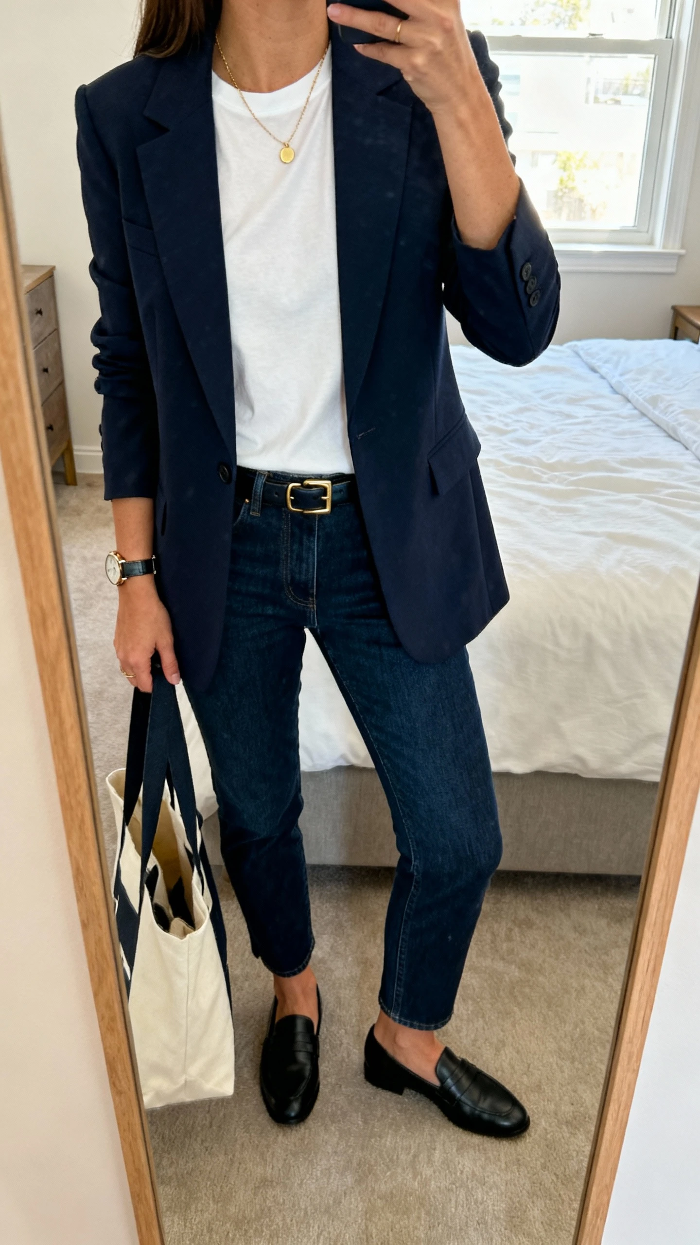 iPhone mirror selfie of a woman wearing a tailored navy blazer over a crisp white tee, dark blue jeans, black loafers, slim leather belt, delicate gold necklace and watch, tote bag on arm, face not visible, cropped at neck, apartment bedroom mirror, morning window light, natural iPhone photo quality.