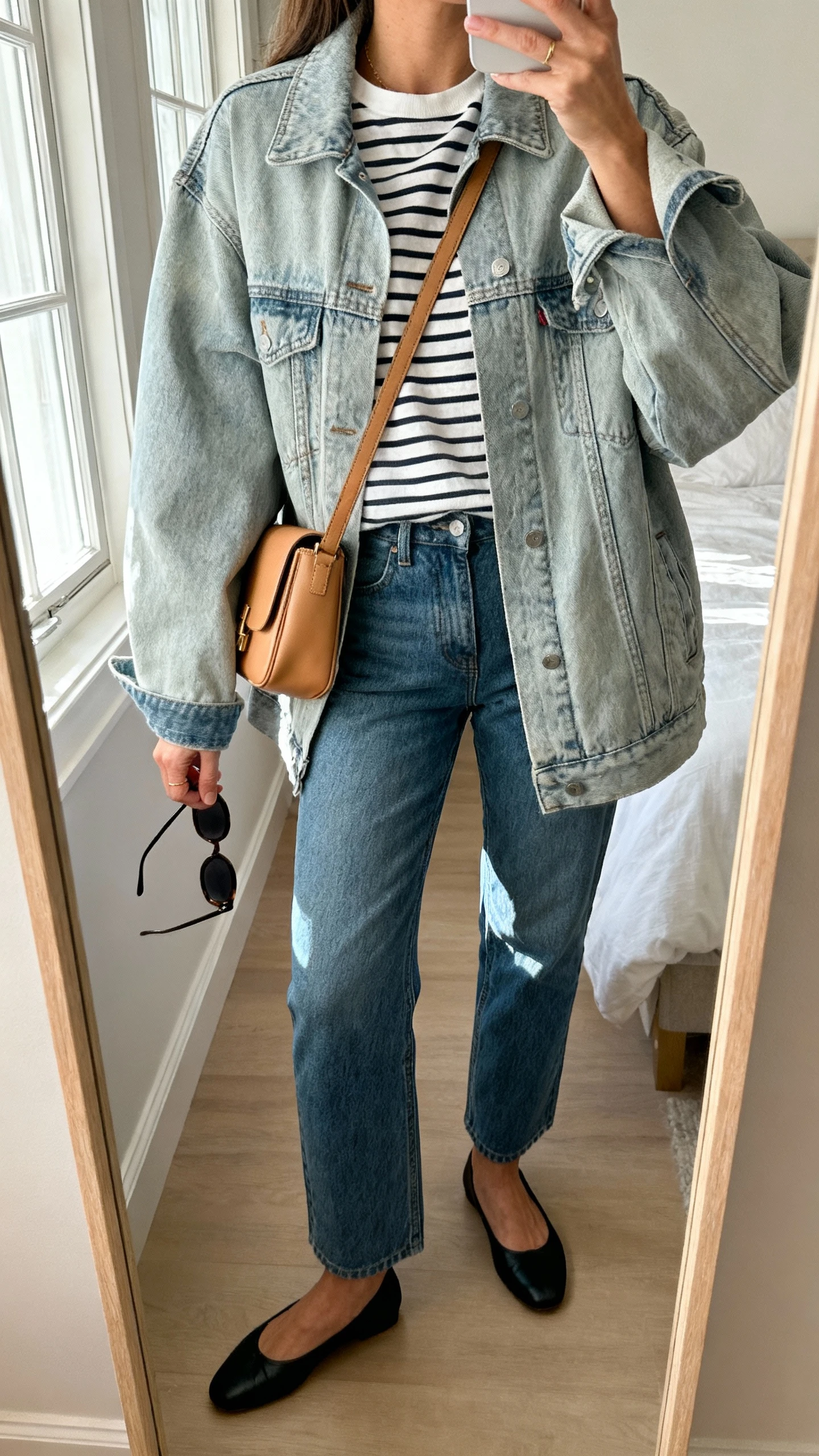 iPhone mirror selfie of a woman wearing an oversized light-wash denim jacket over a classic Breton striped tee, straight-leg blue jeans, black ballet flats, a tan crossbody bag, and slim sunglasses in hand; cropped at neck, bedroom mirror, soft morning window light, face not visible, natural iPhone photo quality.
