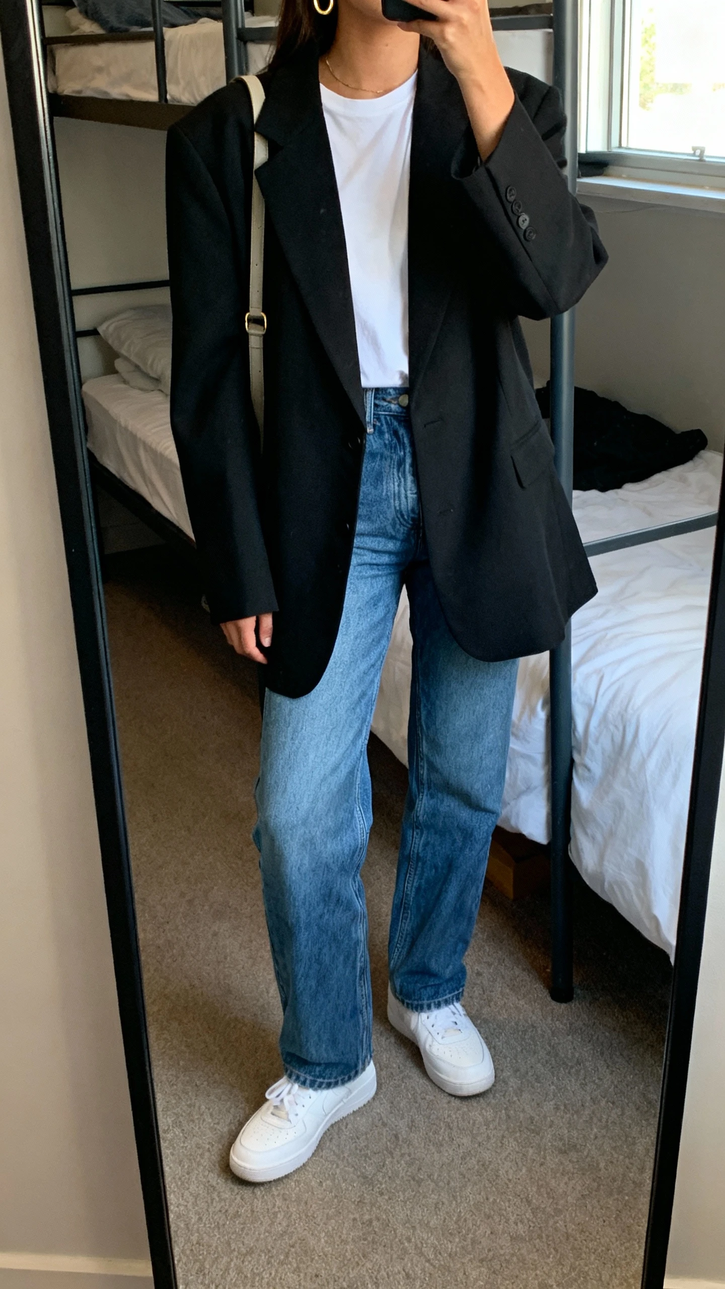 iPhone mirror selfie of a woman wearing an oversized black blazer over a white tee, straight-leg blue jeans, white sneakers, simple gold hoops, shoulder bag, face not visible, cropped at neck, dorm bedroom mirror, natural window light, natural iPhone photo quality.