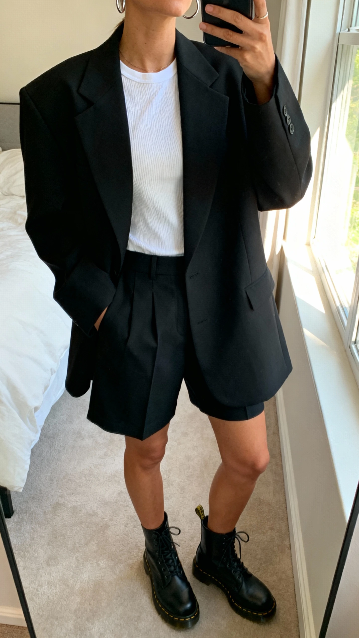 iPhone mirror selfie of a woman wearing an oversized black blazer set (boxy blazer and matching tailored shorts), white ribbed tee, black Doc Martens, simple silver hoops, face not visible, cropped at neck, bedroom mirror with natural window light, natural iPhone photo quality.